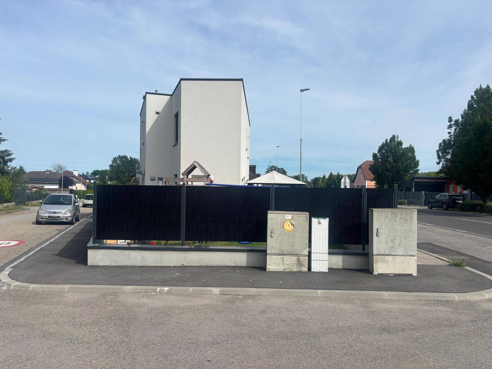 Ansicht des modernen Hauses von der Straßenseite mit dunklem Zaun und Stromkästen.