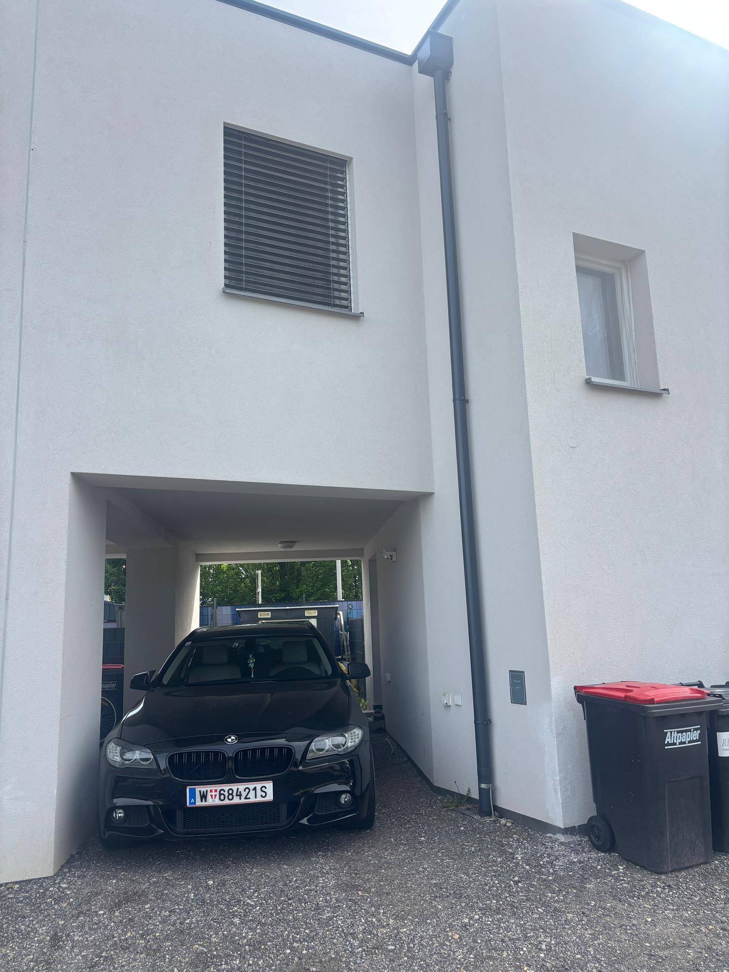 Überdachter Stellplatz für ein Auto unter dem modernen Haus mit Mülltonnen daneben.