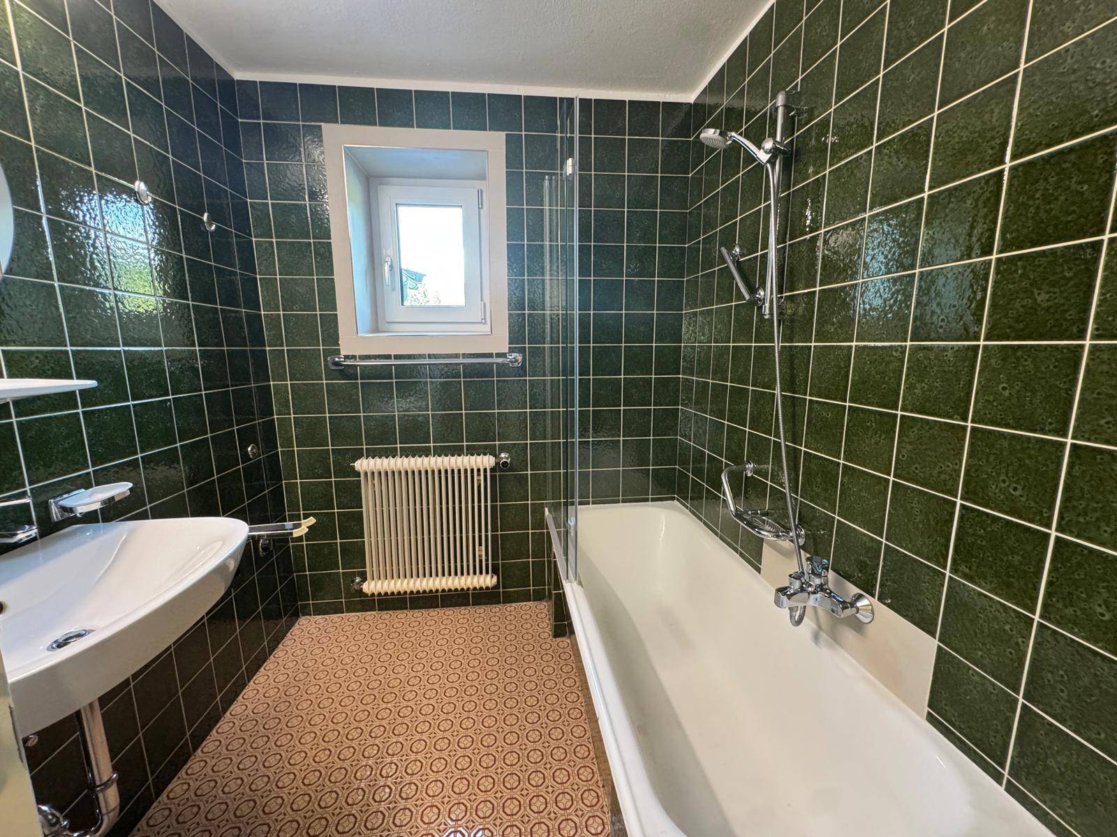 Badezimmer mit dunkelgrünen Wandfliesen, gemustertem Boden und Badewanne mit Duschfunktion.