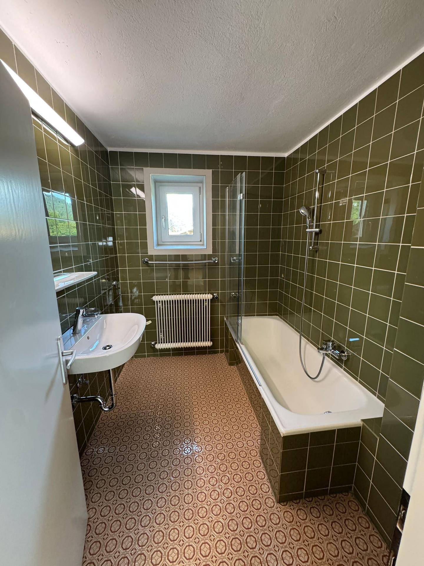 Badezimmer mit grünen Fliesen, Badewanne mit Duschfunktion und Fenster.