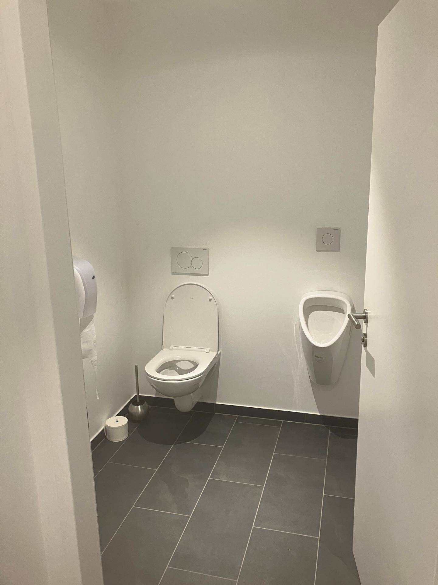 Saubere und moderne Toilette mit Wand-WC, Urinal und dunklen Fliesen auf dem Boden.