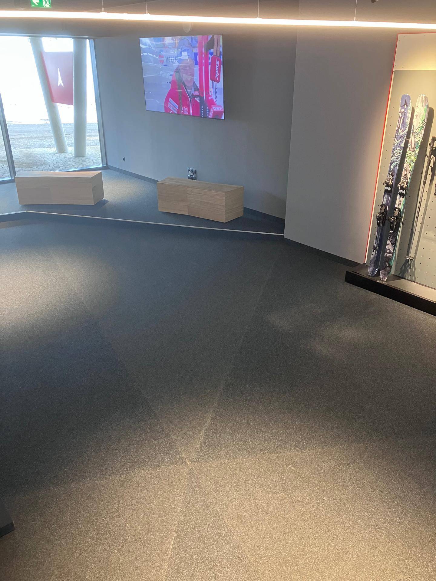 Innenansicht eines Verkaufsraums mit Teppichboden, einem Bildschirm und großen Fenstern im Hintergrund, die Tageslicht hereinlassen.