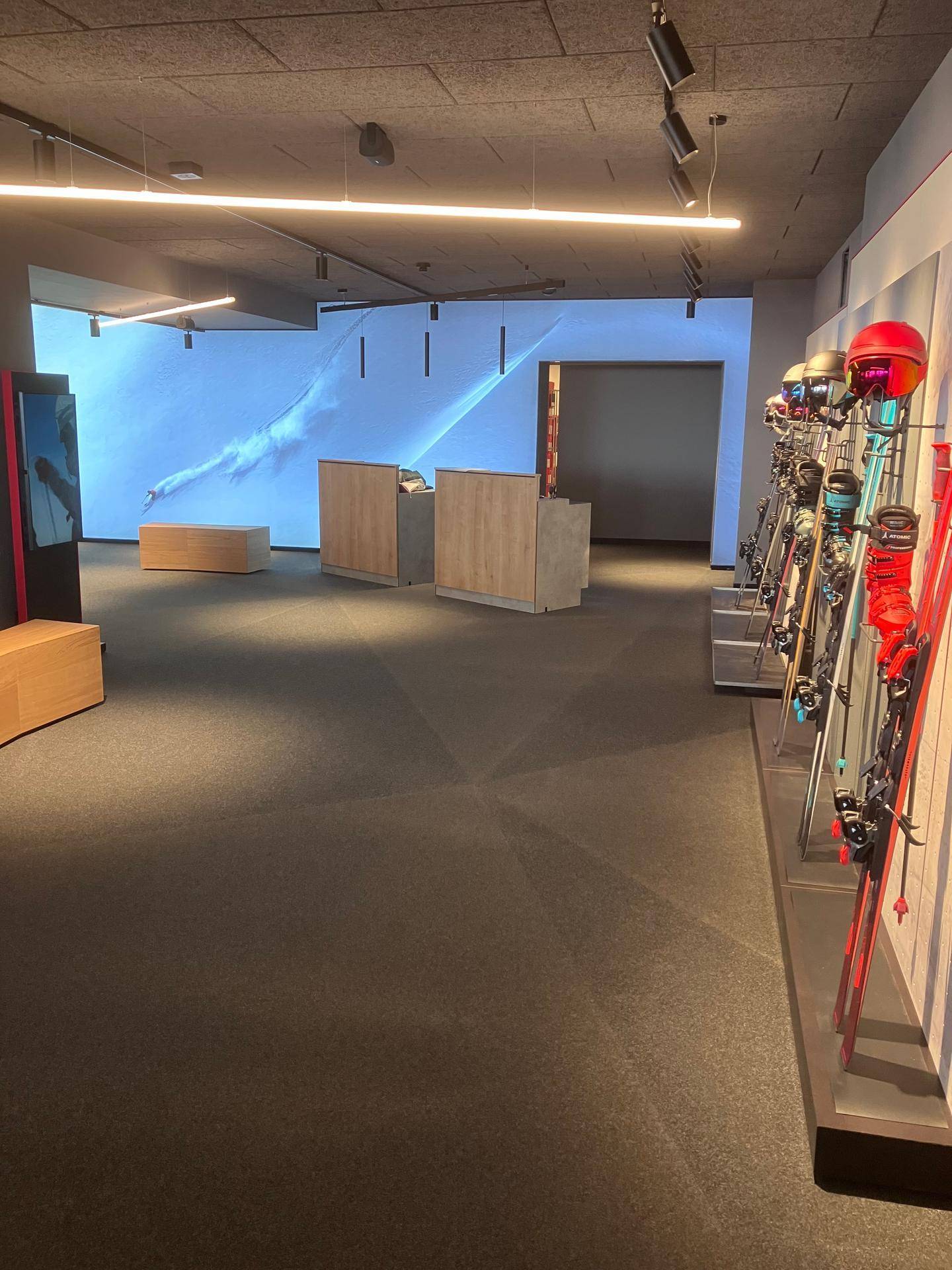 Verkaufsraum mit Skiausrüstung an den Wänden und einer großen Projektionsfläche, die eine Skipiste zeigt.