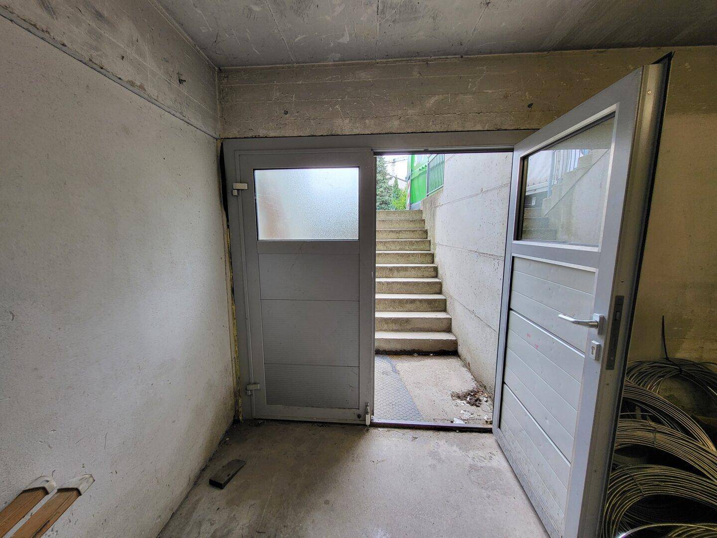 Eingangsbereich im Untergeschoss mit offener Tür, die zu einer Außentreppe aus Beton führt.