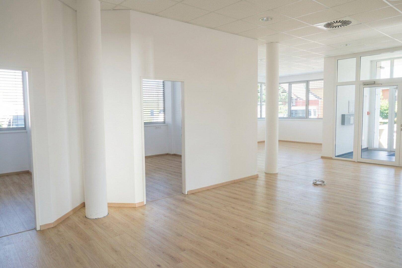 Großzügiger, offener Bürobereich mit vielen Fenstern, Säulen und hellem Laminatboden.