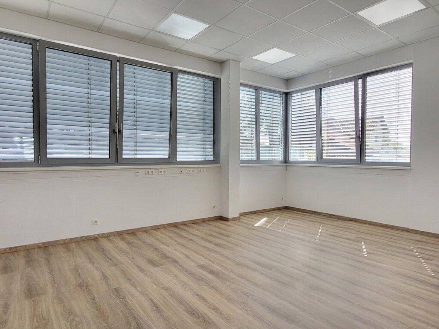 Leeres Büro mit großen Fenstern, Jalousien und hellem Laminatboden, bereit zur individuellen Einrichtung.