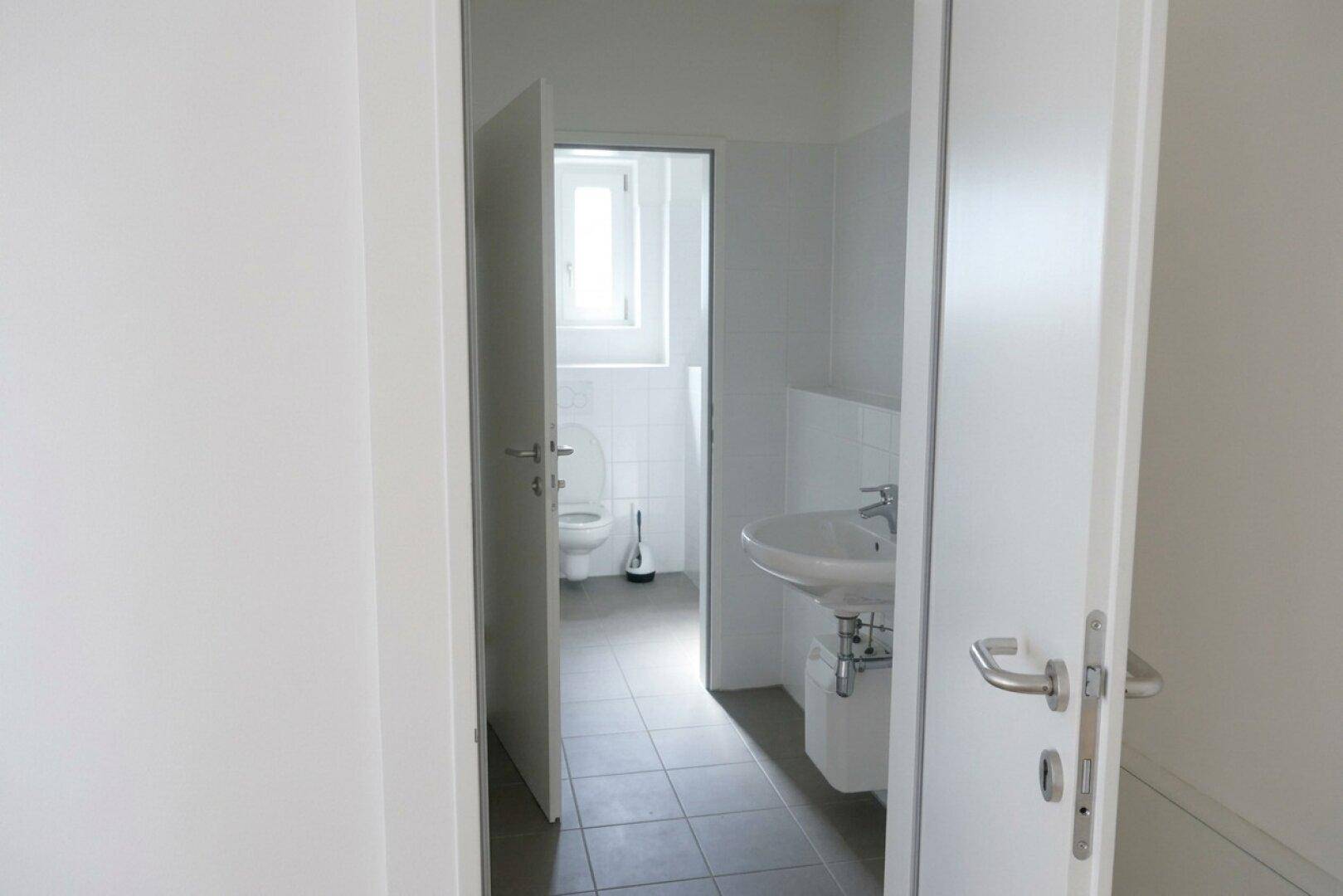 Funktionale Toilette mit Waschbecken, Fenster und grauen Bodenfliesen in einem gepflegten Zustand.