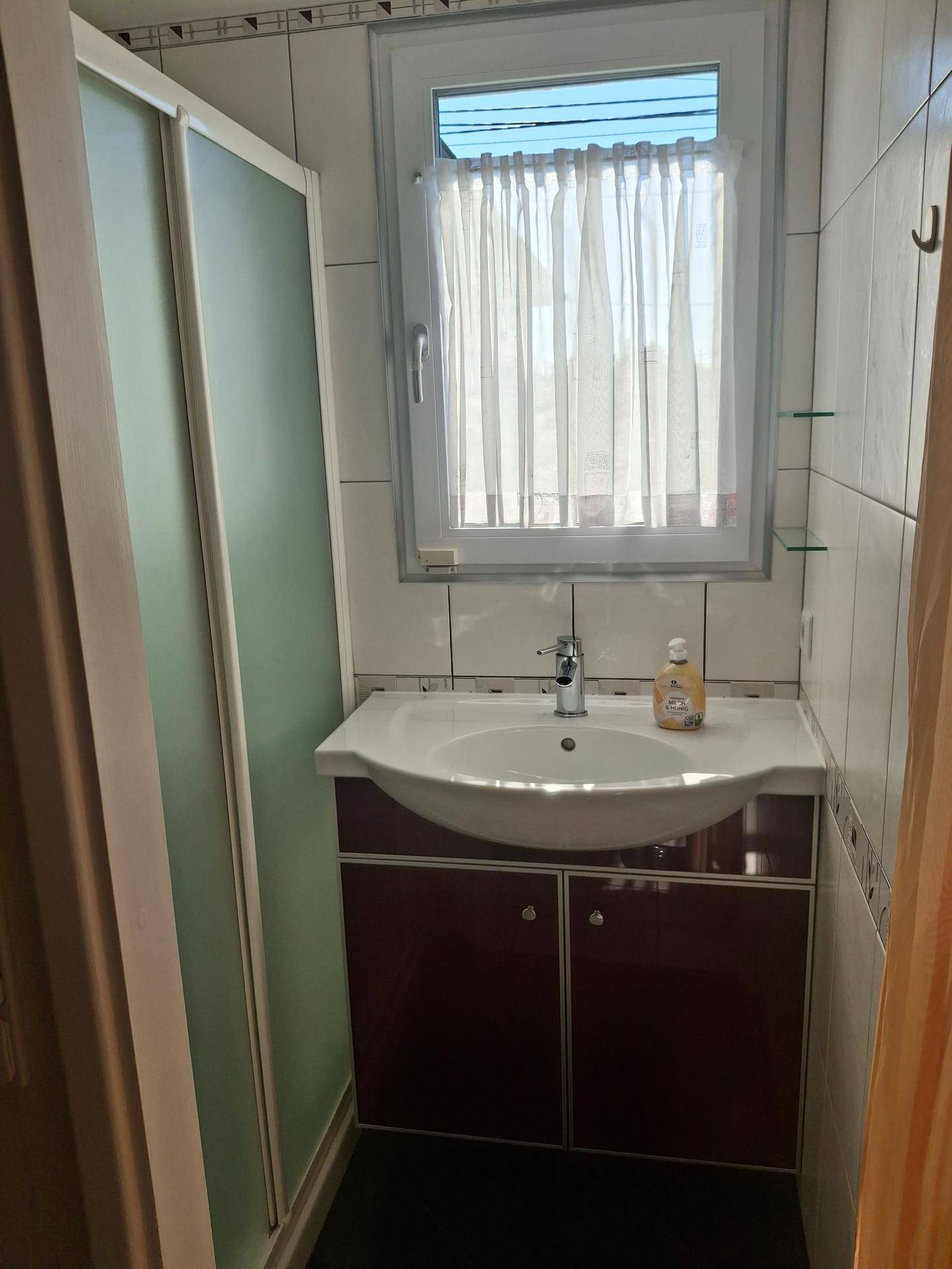 Kleines Badezimmer mit Duschkabine, Waschtischunterschrank und Fenster mit Vorhang.