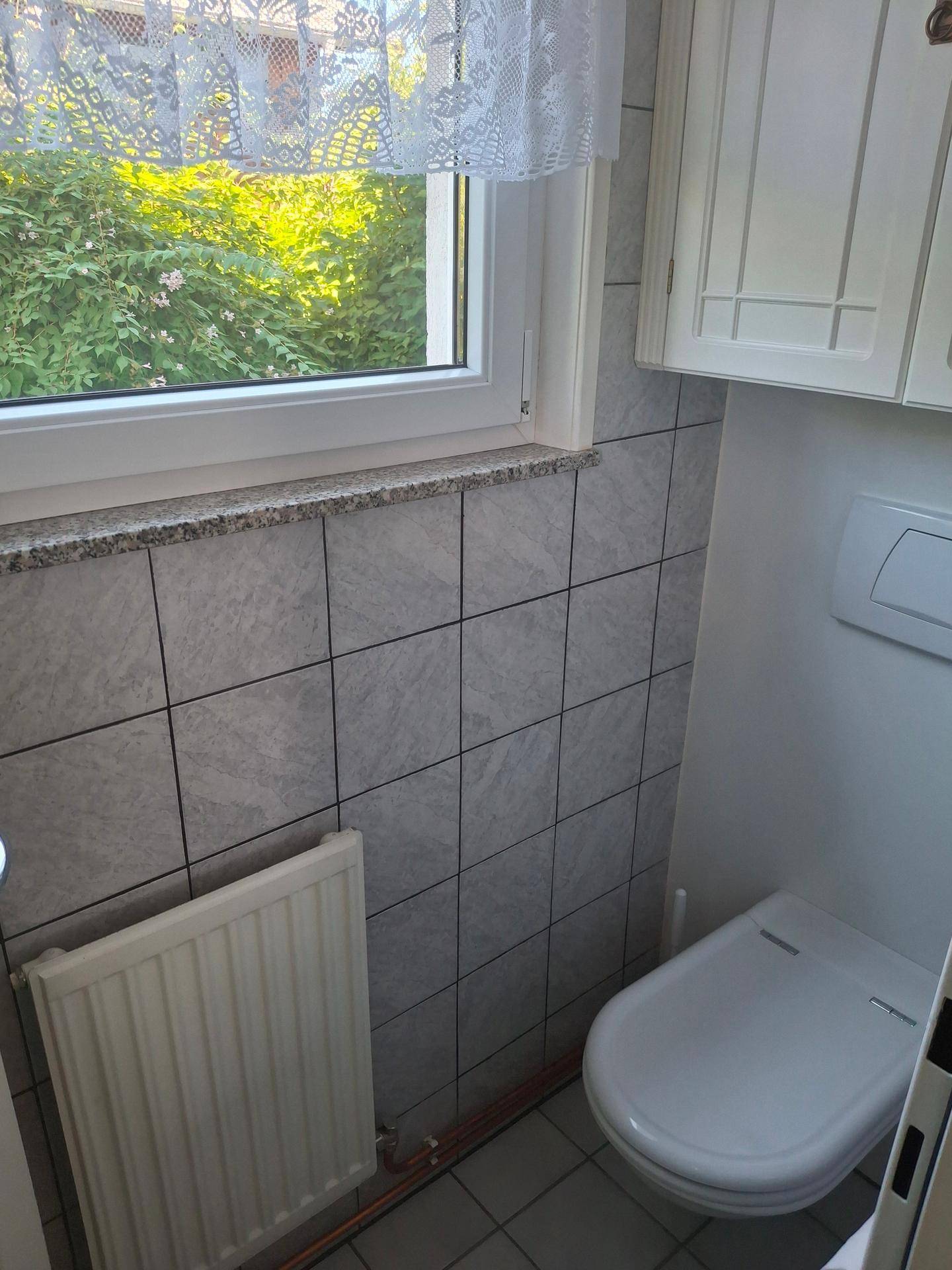 Gäste-WC mit grauen Fliesen, Toilette, Heizkörper und Fenster mit Blick ins Grüne.