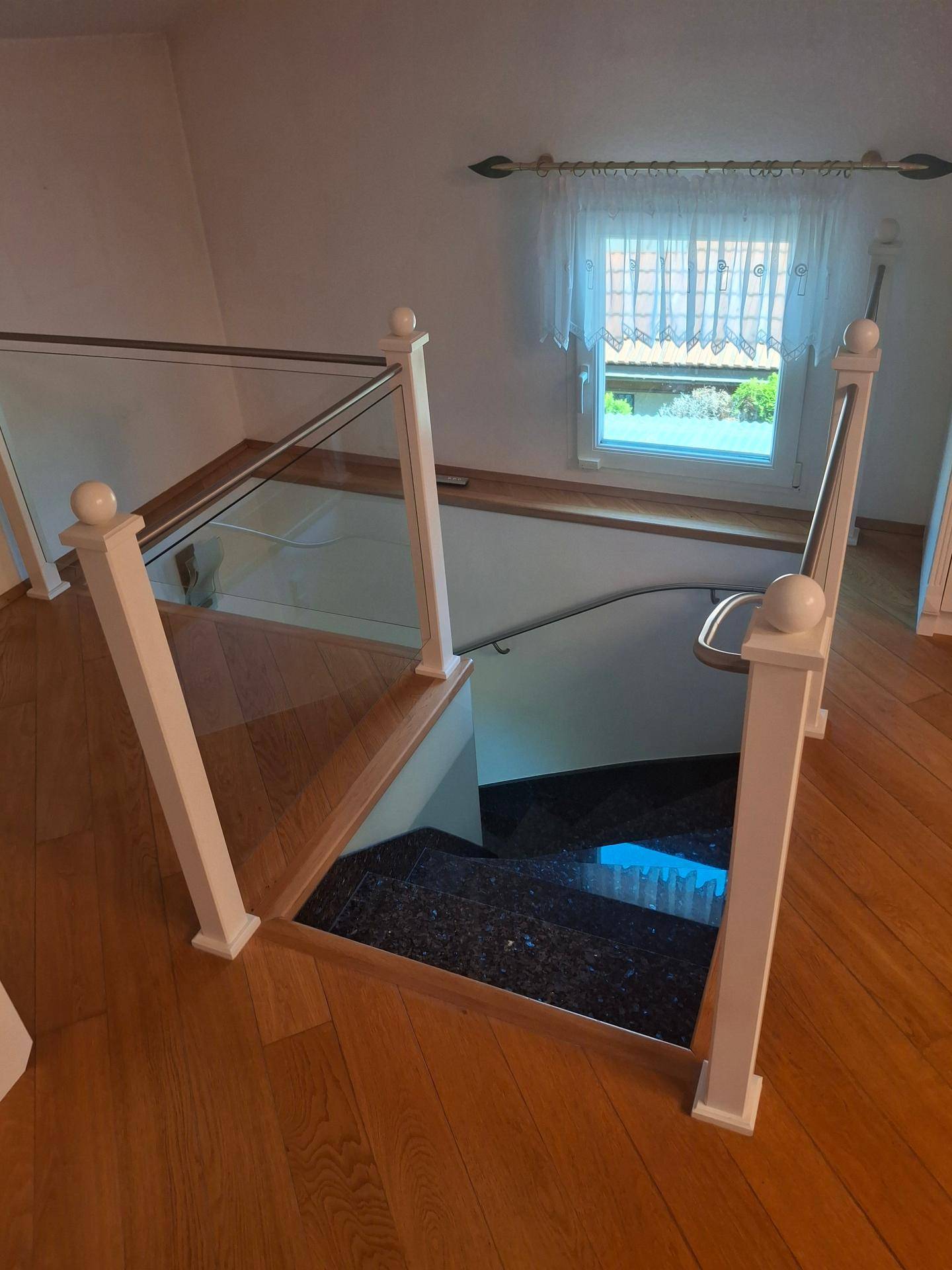 Holztreppe mit Glasgeländer und weißem Holzpfosten, die nach unten führt, mit Fensterblick.