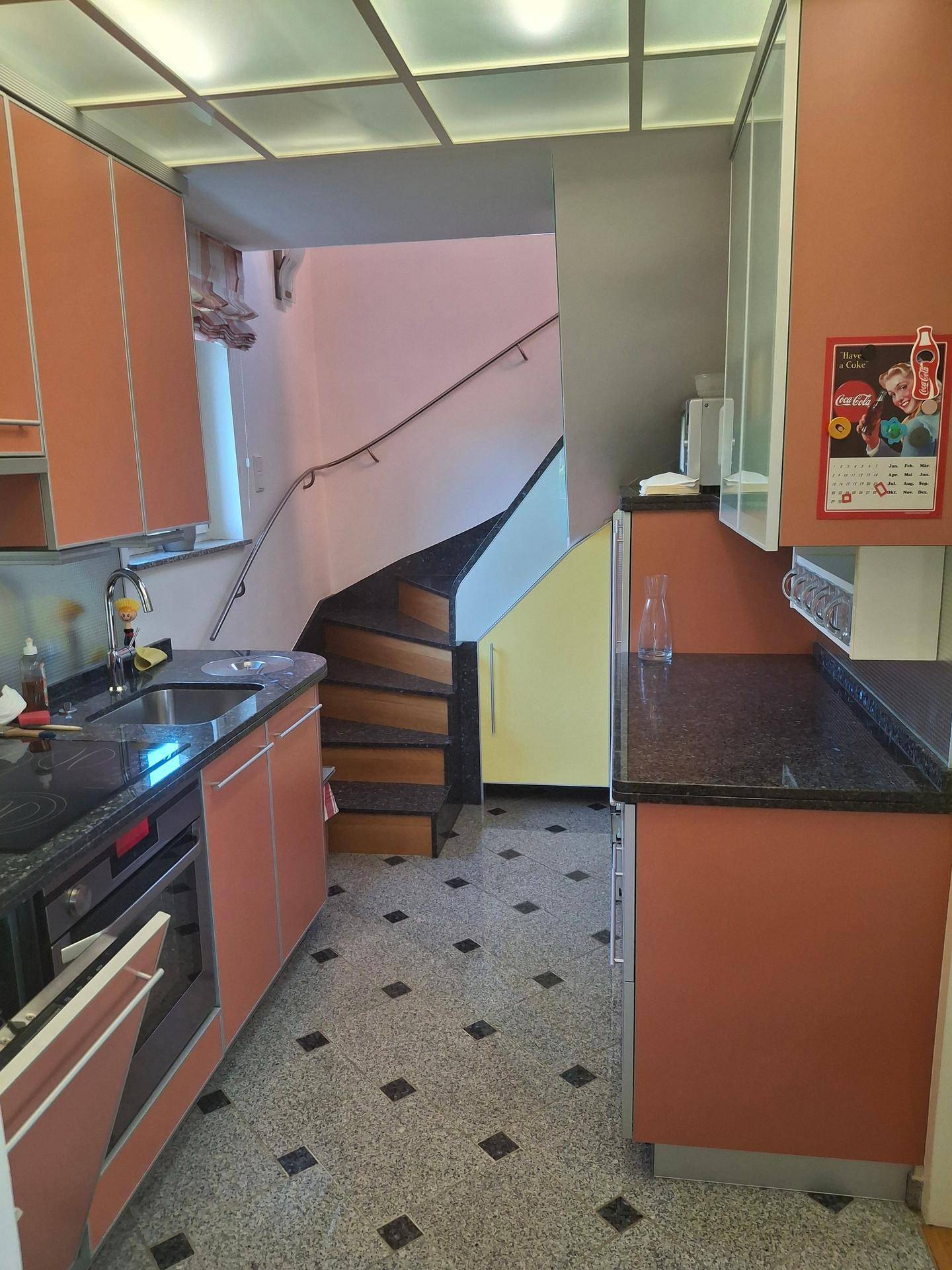 Küche mit orangefarbenen Schränken und dunkler Arbeitsplatte, im Hintergrund eine Wendeltreppe.