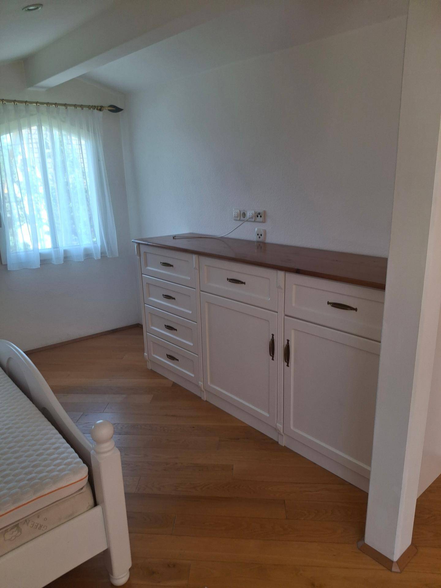 Schlafzimmer mit maßgefertigten weißen Schränken, Holzplatte und Fenster mit Vorhängen.