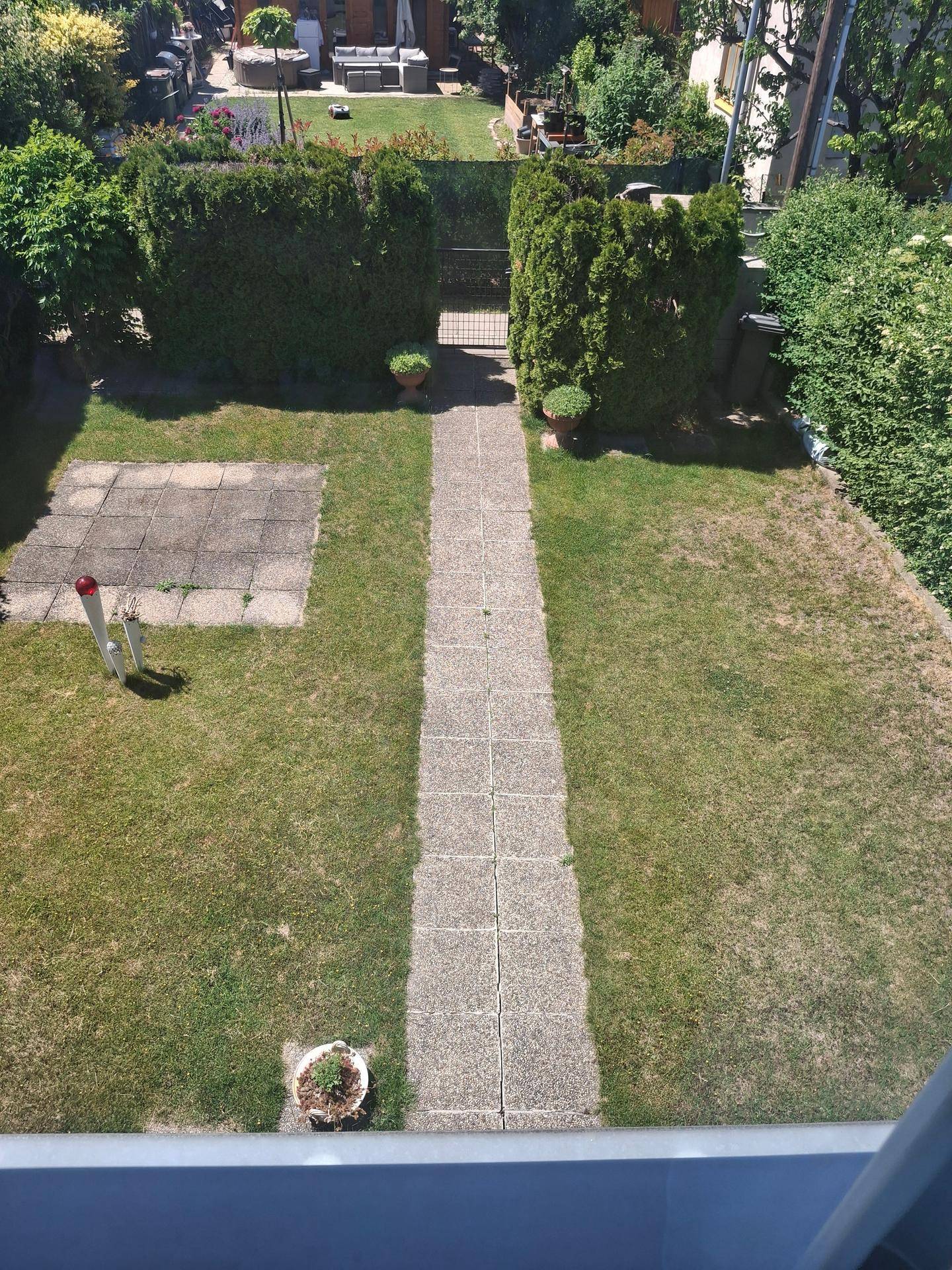 Blick von oben auf einen gepflegten Garten mit Rasenfläche, Steinweg und hohen Hecken.
