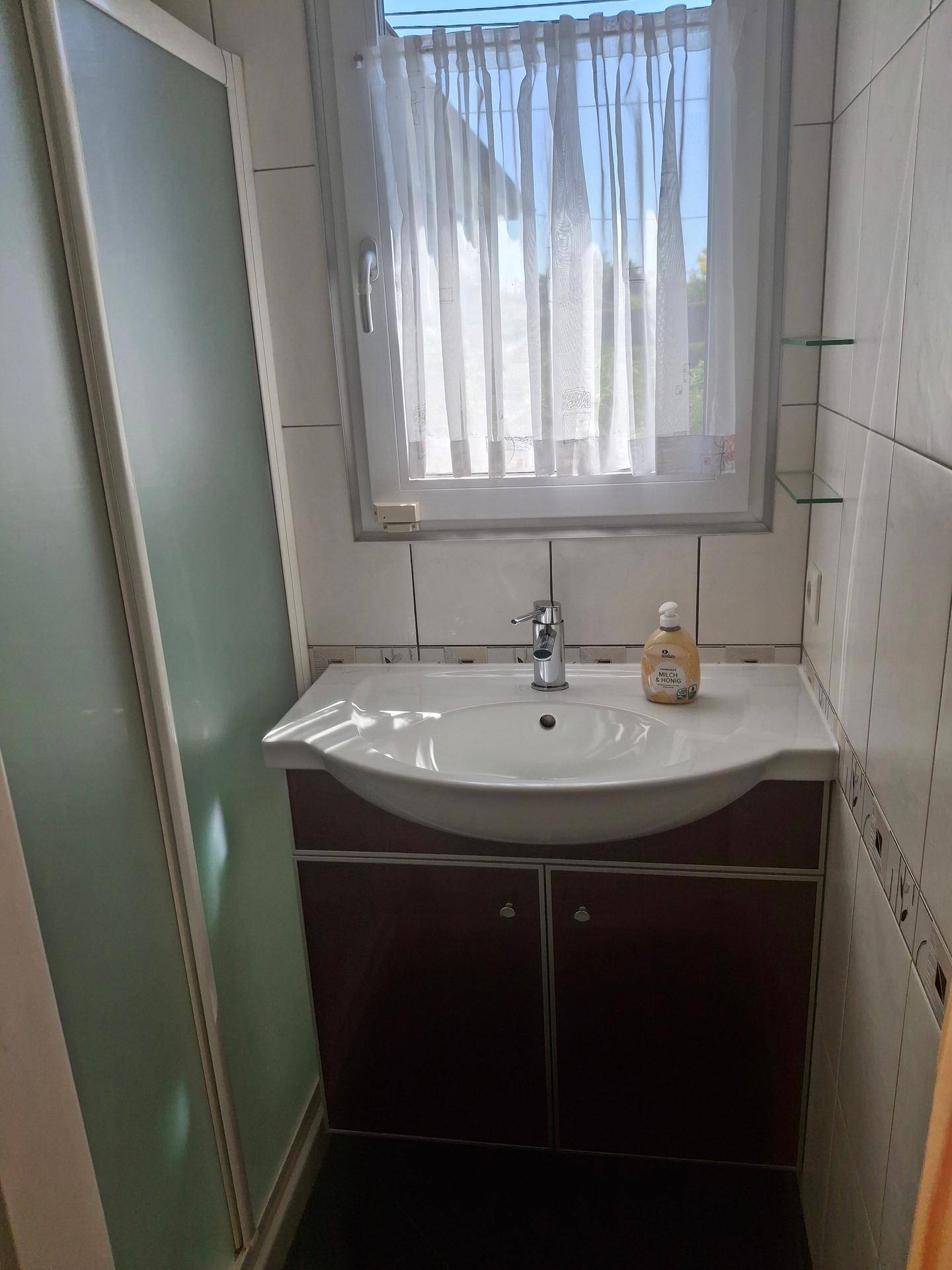 Kleines Badezimmer mit Duschkabine, Waschtischunterschrank und Fenster mit Vorhang.