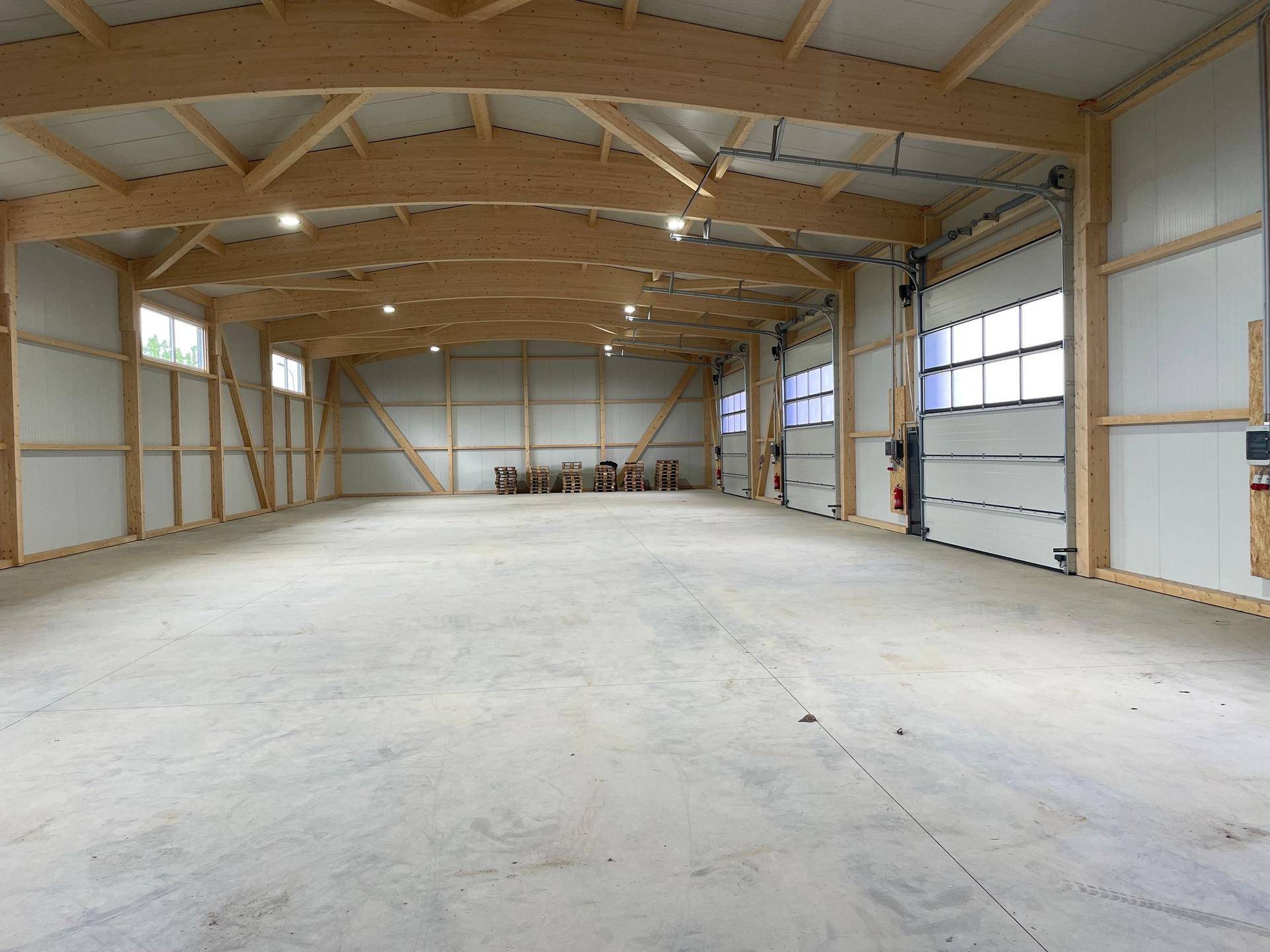 Geräumige, leere Lagerhalle mit hohen Decken, sichtbaren Holzträgern und großen Rolltoren mit Fenstern.