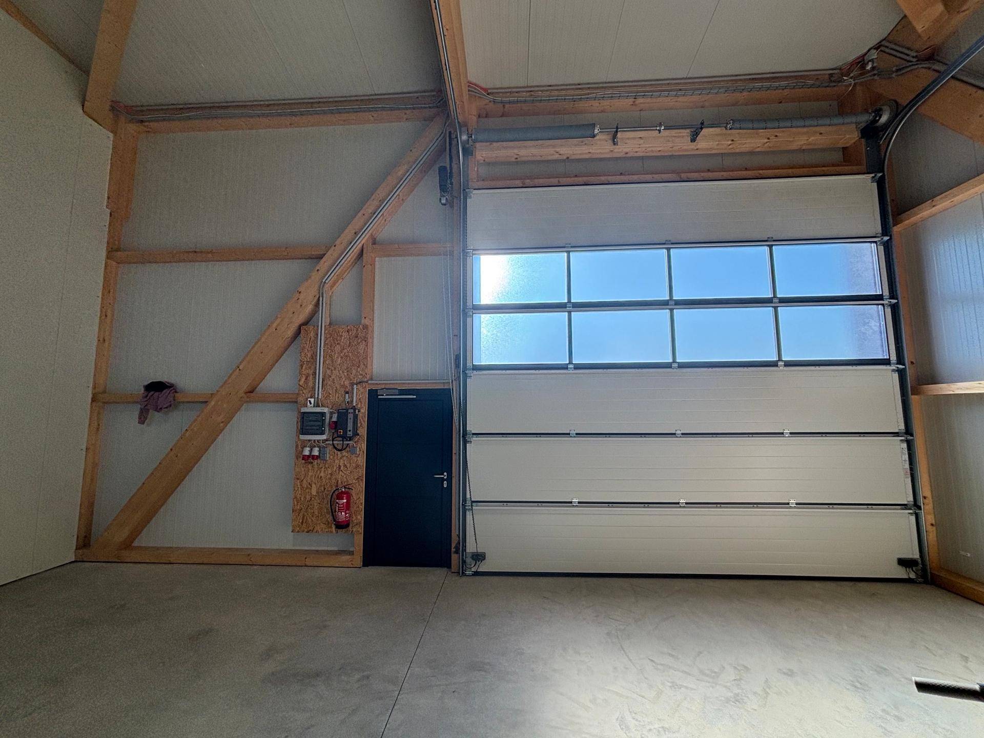 Innenansicht einer Lagerhalle mit Rolltor, Fenstern, Seitentür und sichtbarer Elektroinstallation.