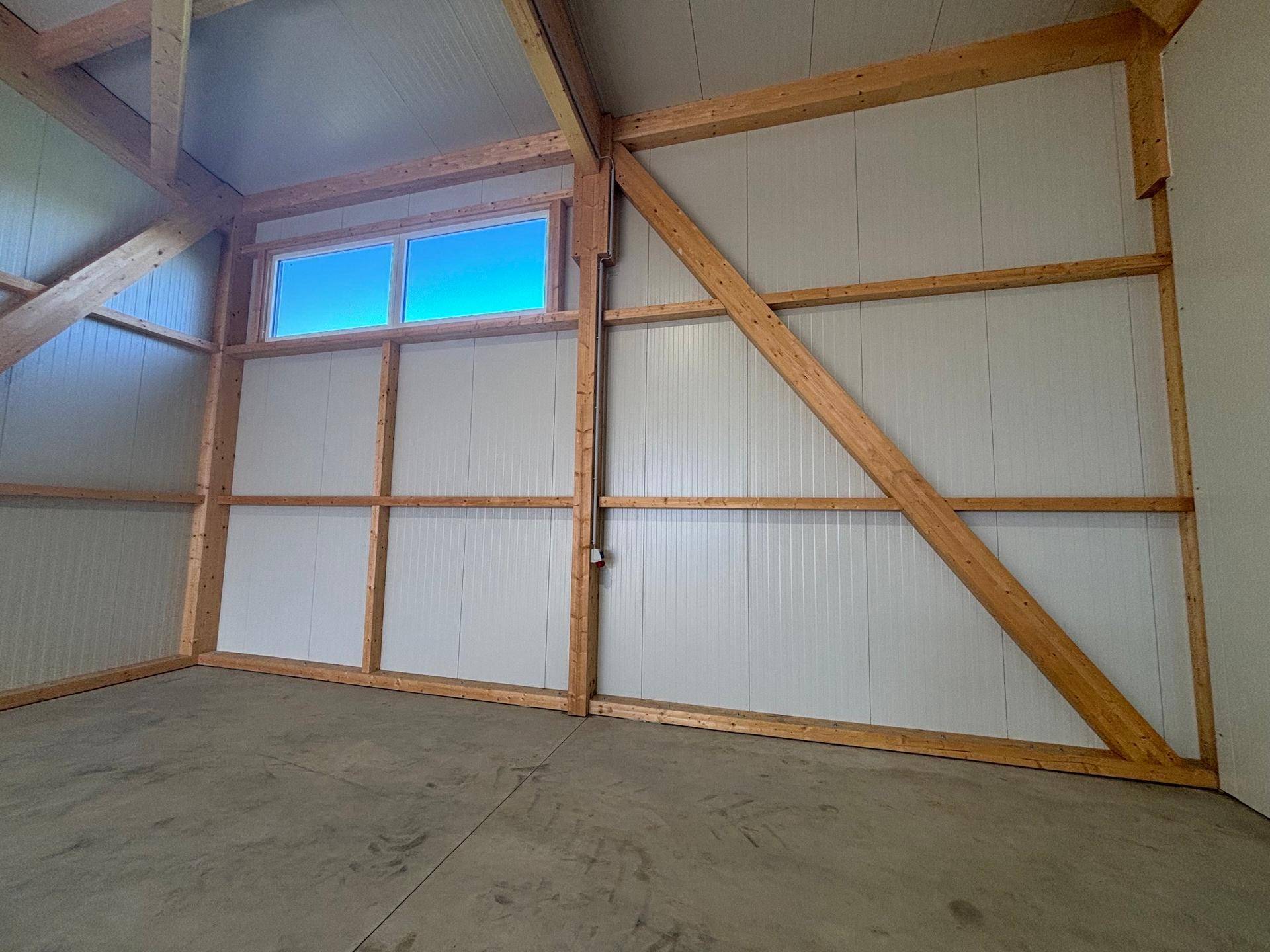 Ecke einer Lagerhalle mit sichtbaren Holzträgern, weißen Wänden und einem hochgelegenen Fenster mit blauem Himmel.