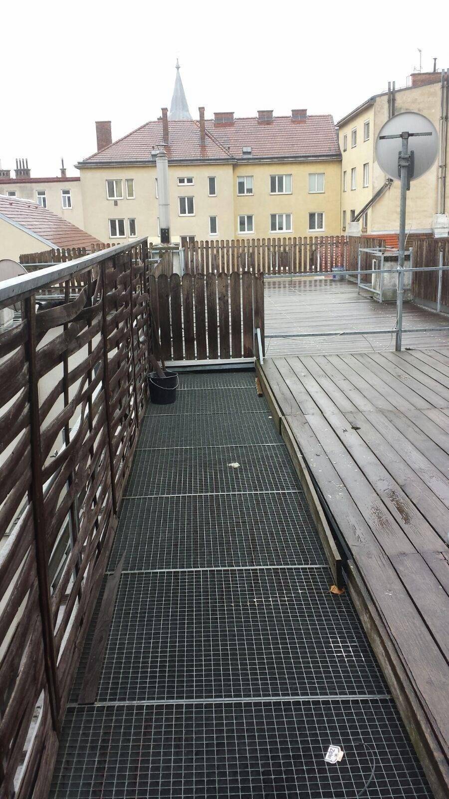 Dachterrasse mit Holzboden und Metallgitter, umgeben von Gebäuden mit Blick auf die Umgebung.