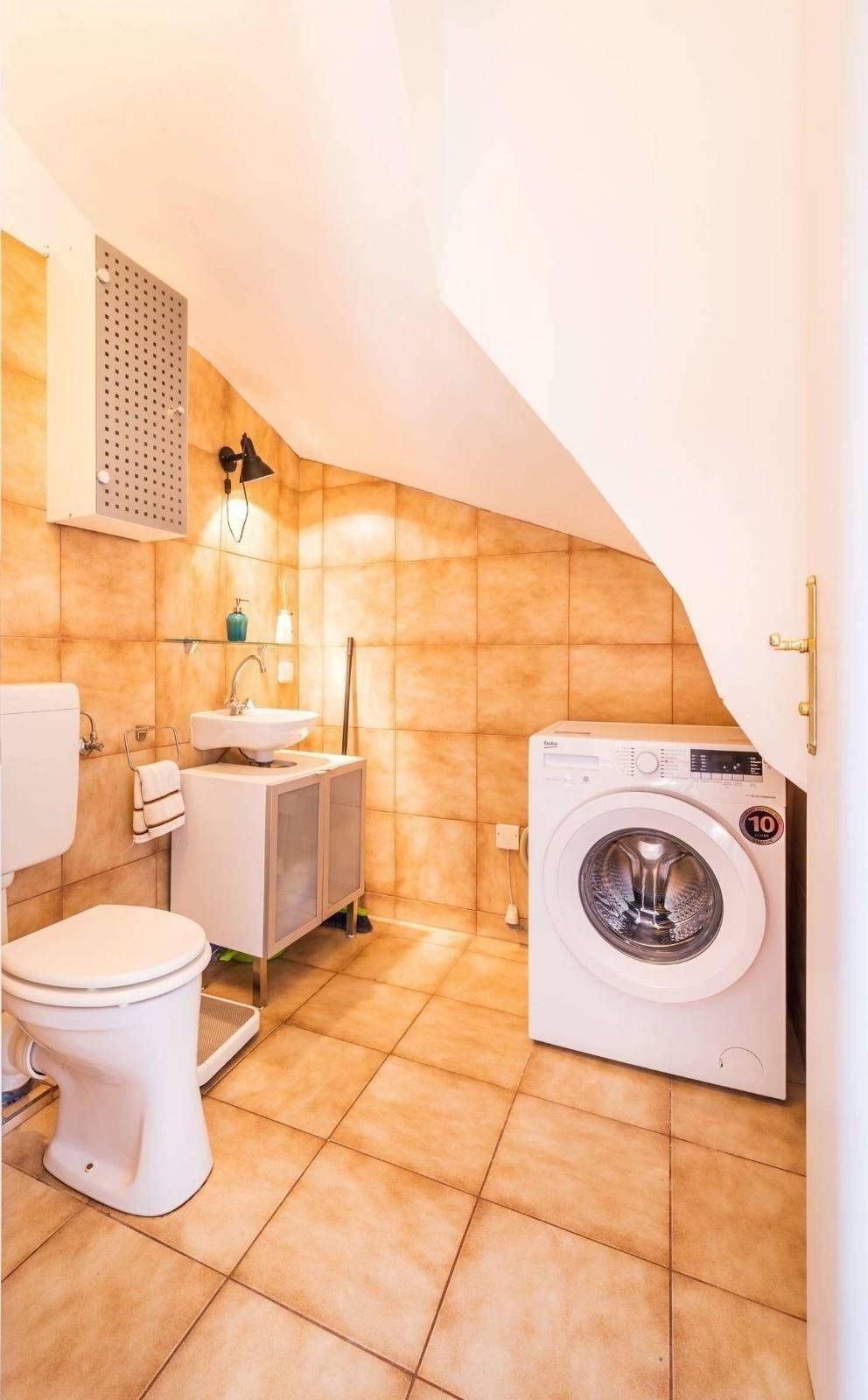 Badezimmer mit Waschmaschine, Toilette und Waschbecken, ausgestattet mit beigefarbenen Fliesen.