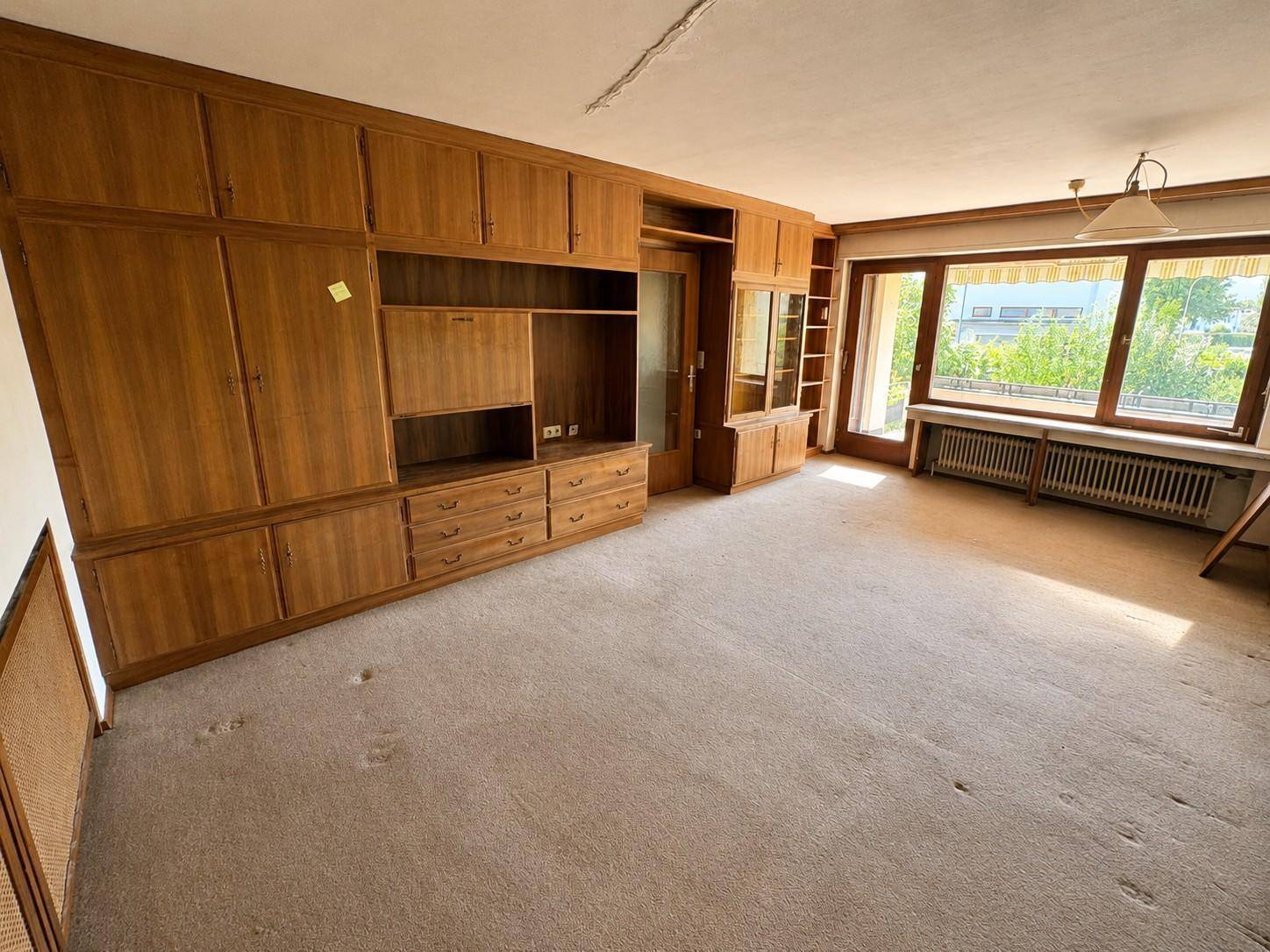 Geräumiges Wohnzimmer mit Teppichboden und großer Holzschrankwand, hell durch Fenster.