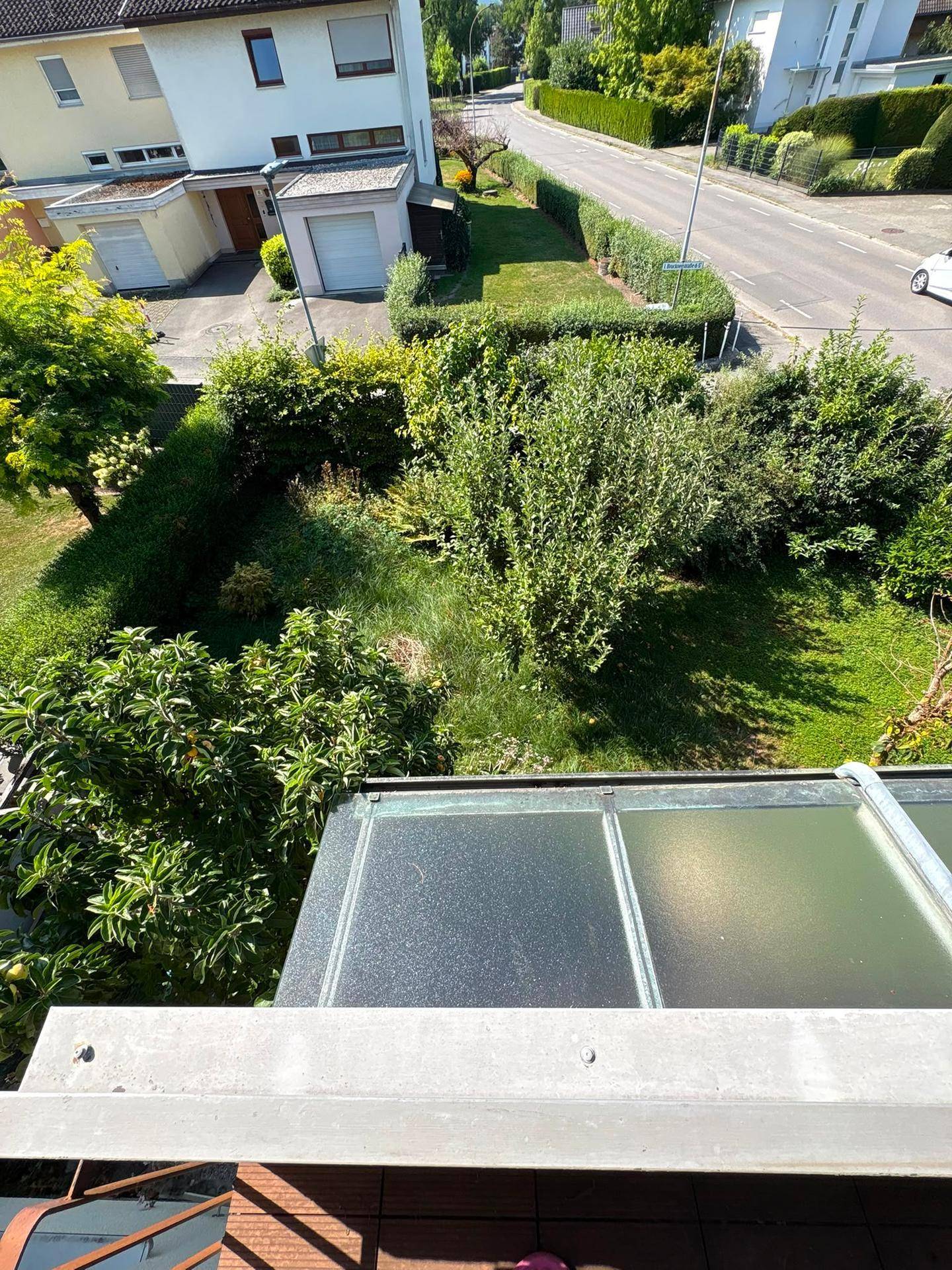 Blick vom Balkon auf einen gepflegten Garten und die dahinterliegende Straße.