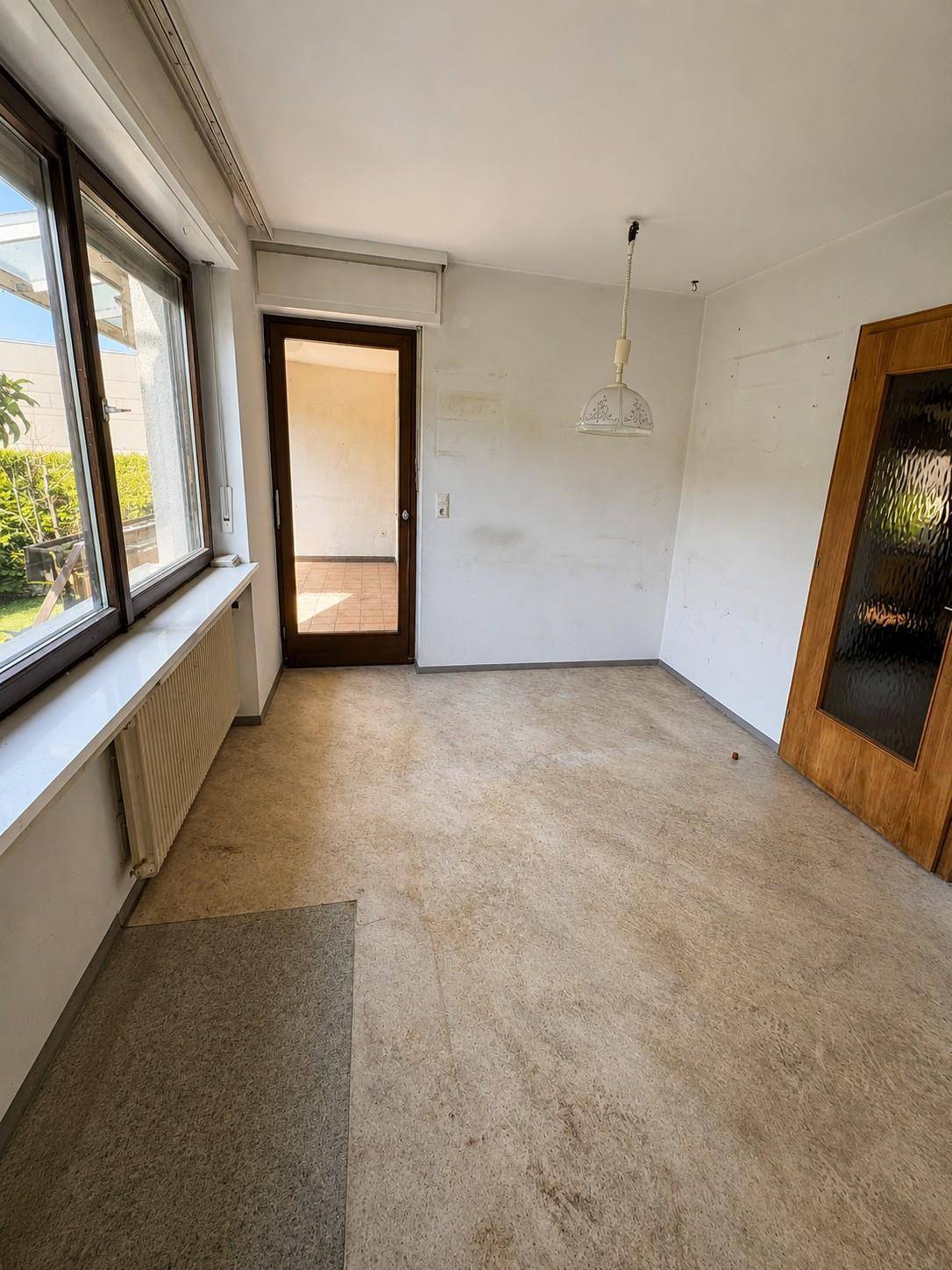 Leerer Raum mit großem Fenster, Zugang nach draußen und gemischtem Bodenbelag.