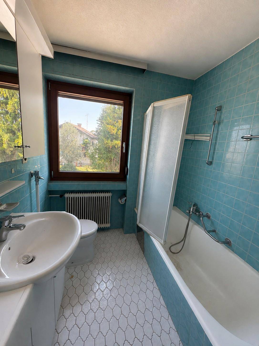 Badezimmer mit blauen Fliesen, Badewanne mit Duschwand, Waschbecken und Fenster.