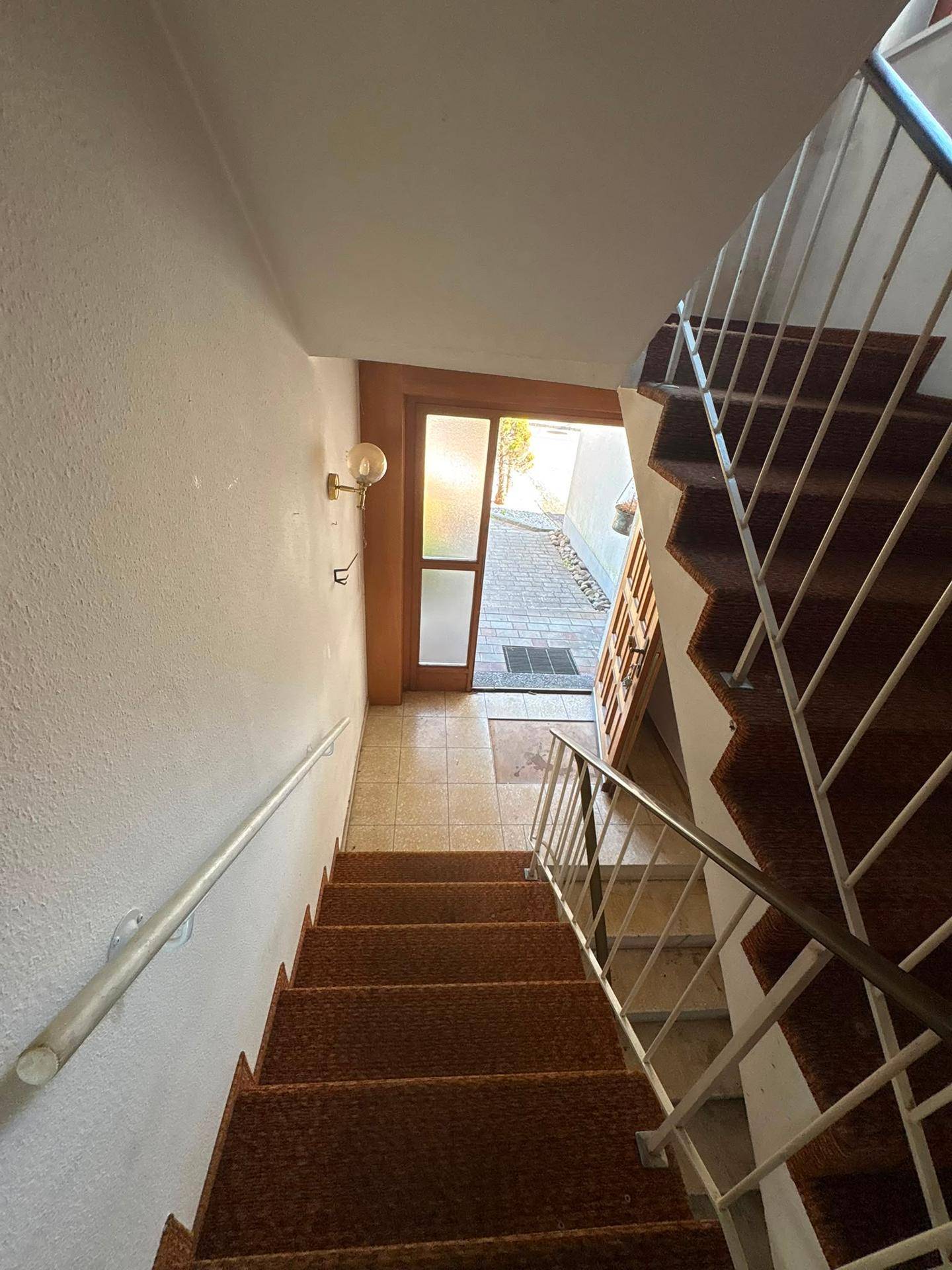 Treppenhaus mit Teppichstufen und Metallgeländer, führt zum hellen Eingangsbereich des Hauses.
