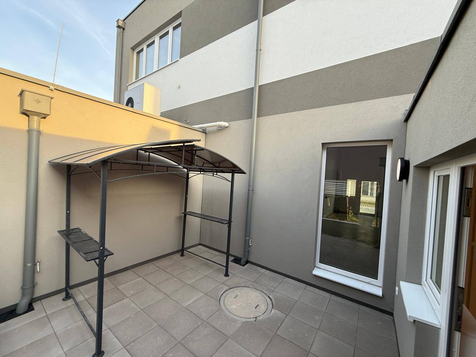 Kleine, gepflasterte Terrasse mit einer Metallüberdachung und Blick auf die Gebäudefassade.
