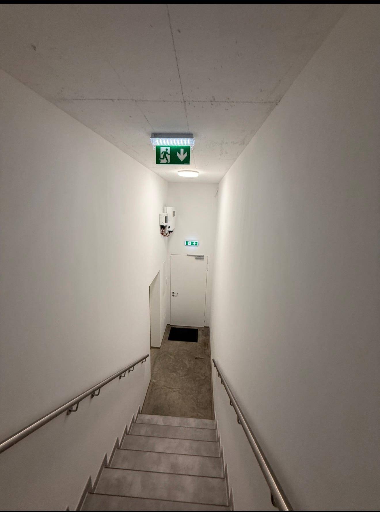 Treppenhaus mit Betonstufen und Handläufen, führt nach unten, mit Notausgangsschild.