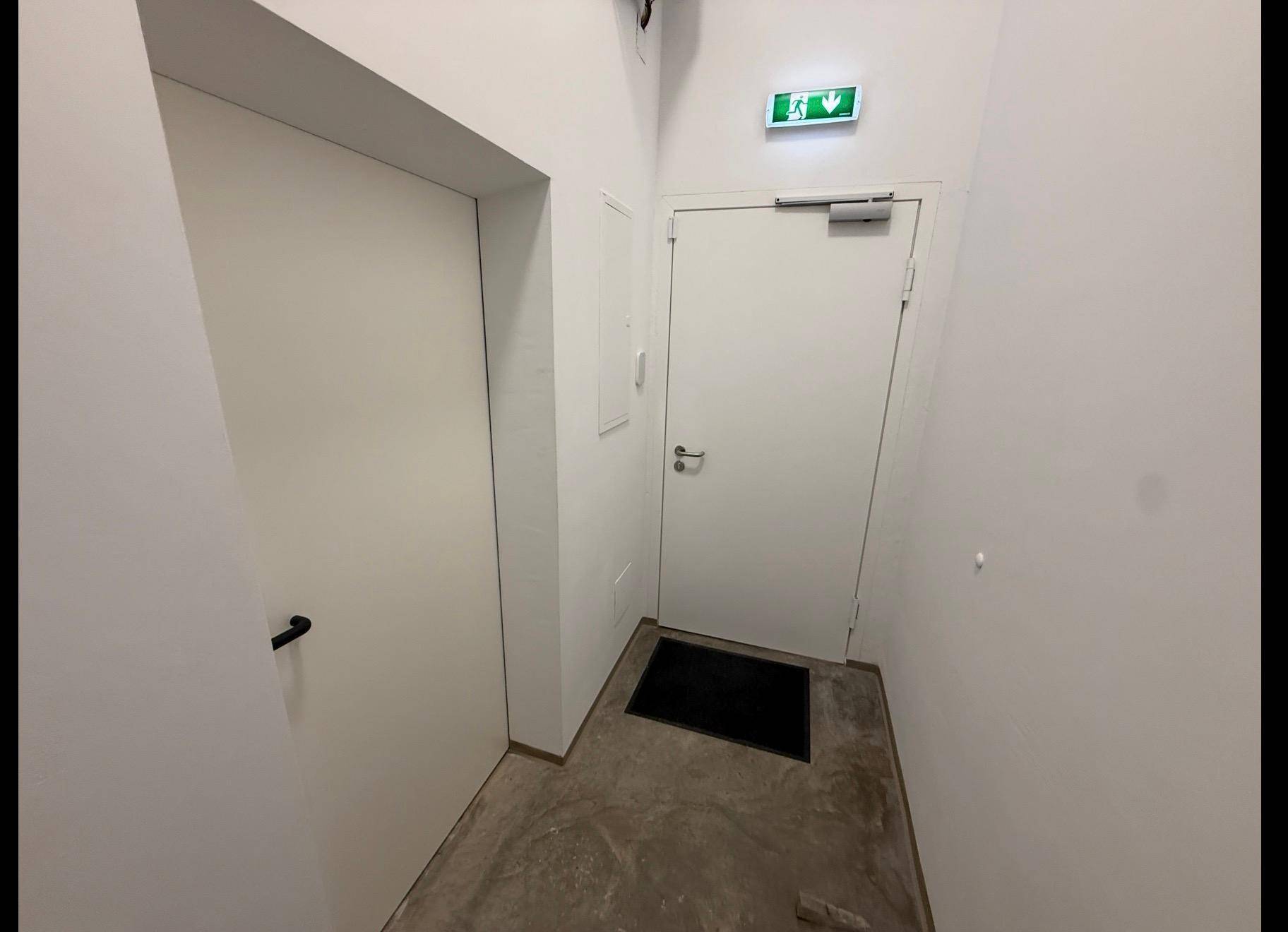 Heller Flur mit zwei Türen und einem grünen Notausgangsschild an der Decke.