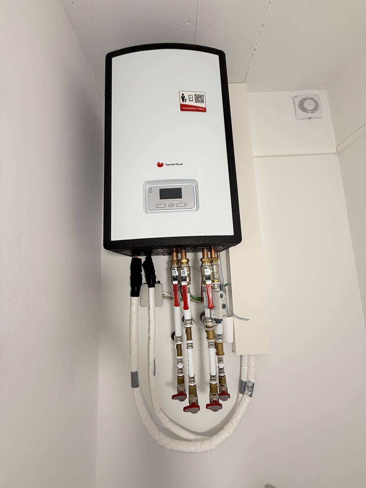 Nahaufnahme eines modernen Warmwasserbereiters mit sichtbaren Anschlüssen an einer weißen Wand.