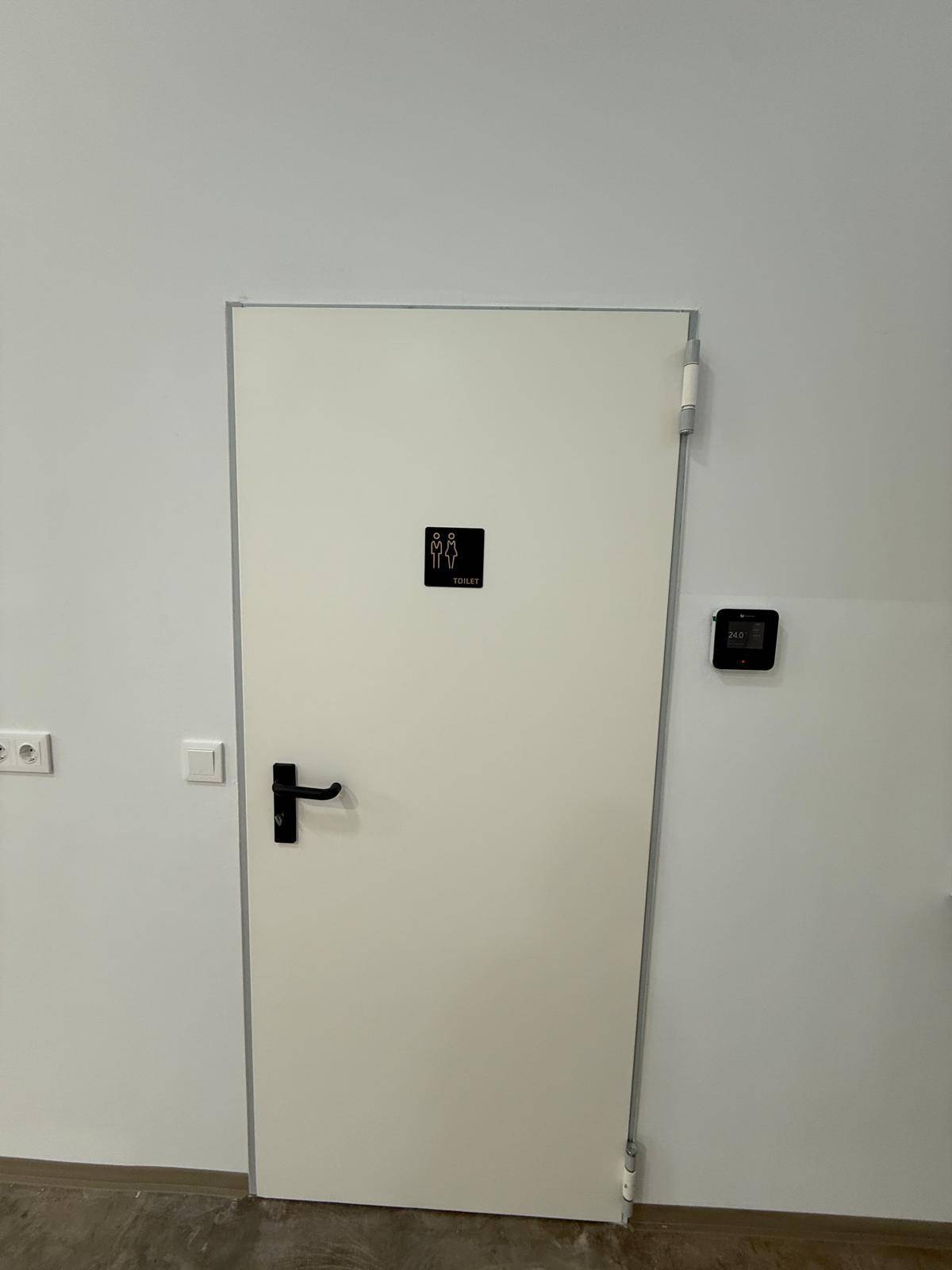 Weiße Tür mit Toilettenschild in einem Flur, daneben ein Thermostat an der Wand.
