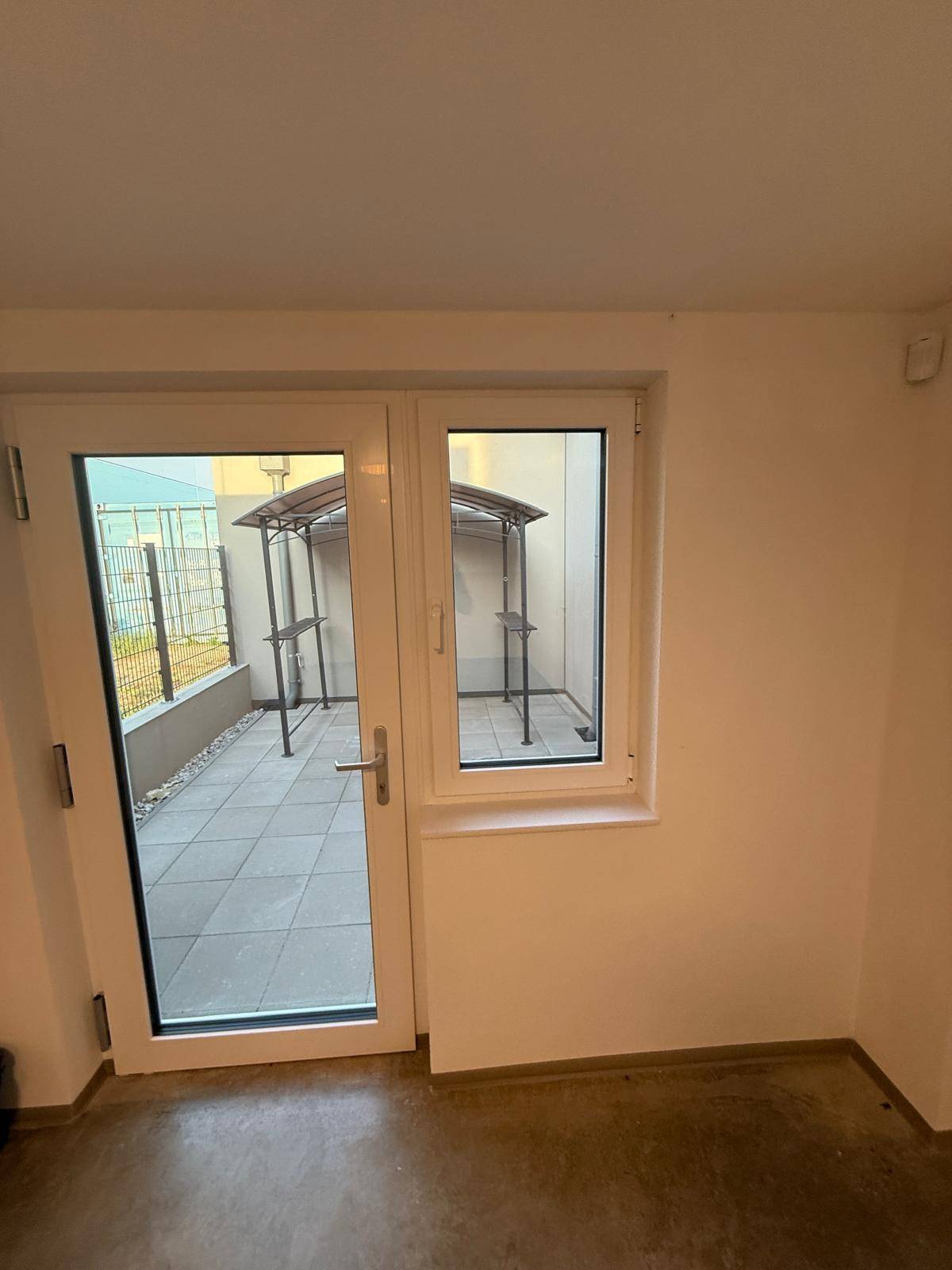 Blick vom Innenraum durch eine Glastür auf eine kleine gepflasterte Terrasse mit Überdachung.
