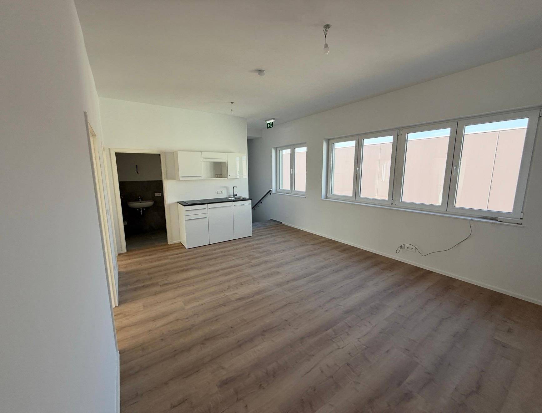 Heller Büroraum mit Laminatboden, großen Fenstern und einer integrierten Küchenzeile.