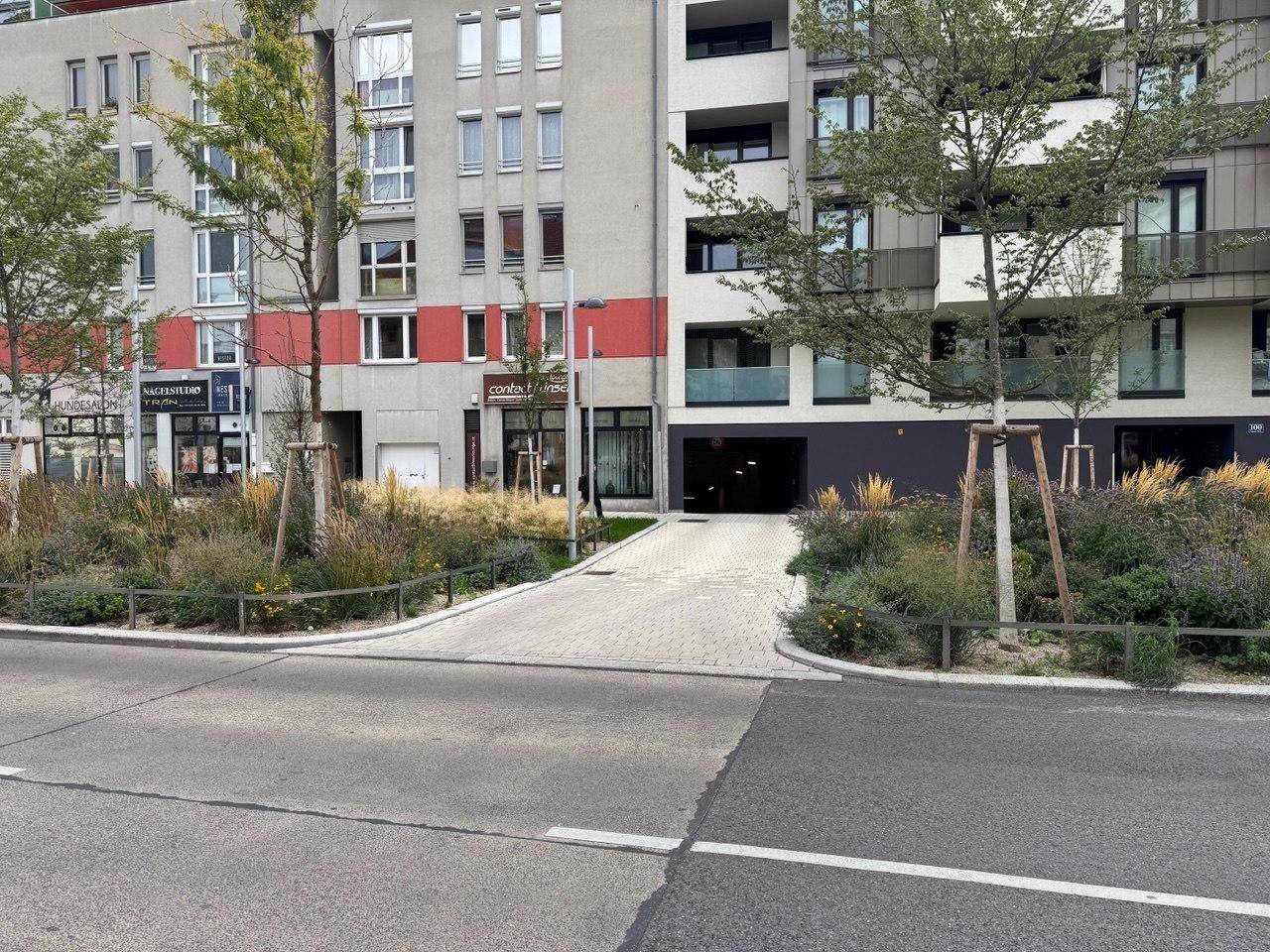Außenansicht eines modernen Gebäudekomplexes mit Zufahrt zur Tiefgarage und gepflegter Bepflanzung.