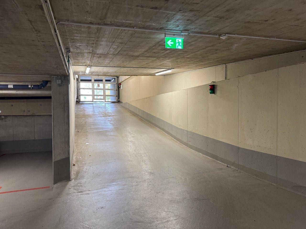 Ausfahrt einer Tiefgarage mit Rampe, Notausgangsschild und Rolltor am Ende.
