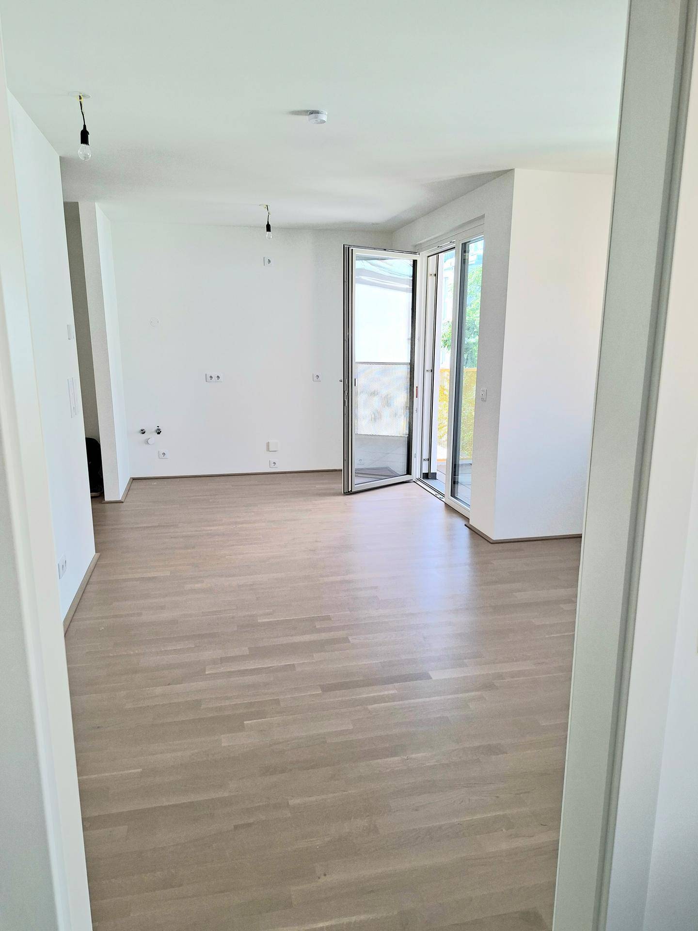 Leerer Raum mit hellem Parkettboden und Zugang zu einem Balkon oder einer Terrasse.