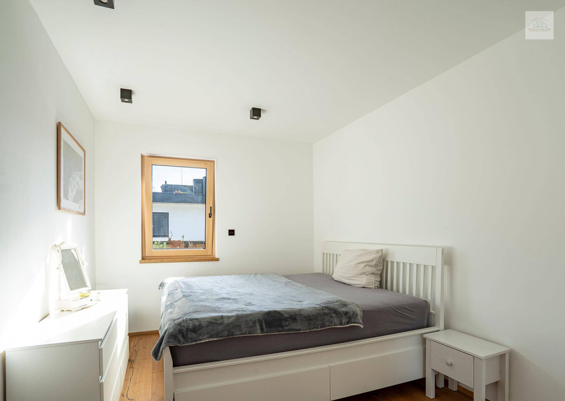 Helles Schlafzimmer mit weißem Bett, Kommode und Holzboden, gut beleuchtet durch ein Fenster.