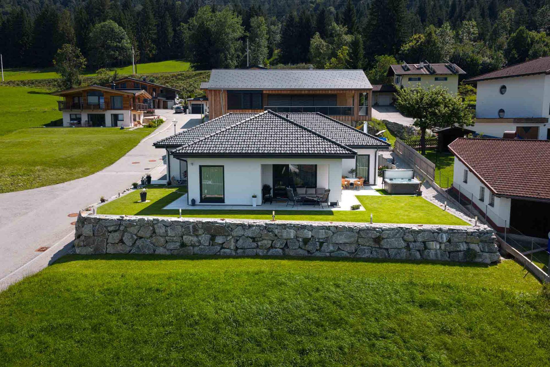 Luftaufnahme des modernen Hauses mit gepflegtem Garten, Steinmauer und Whirlpool in alpiner Umgebung.