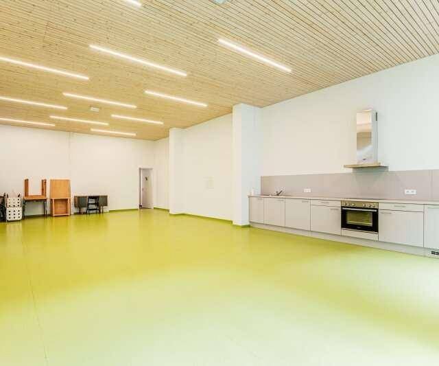 Großer Mehrzweckraum mit heller Holzdecke, leuchtend grünem Boden und kleiner Küchenzeile.