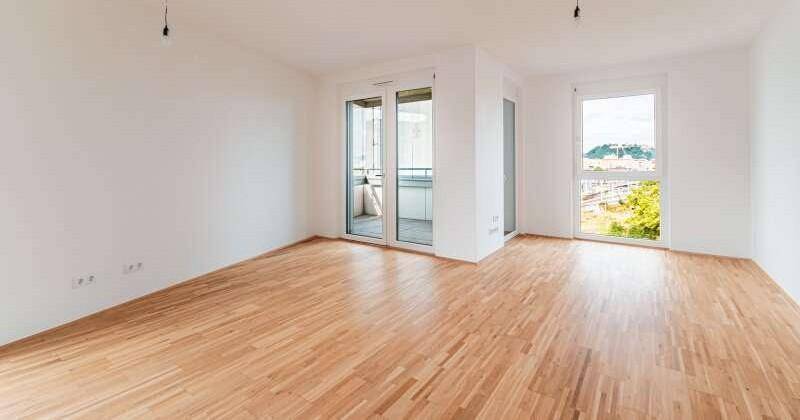 Heller, leerer Wohnraum mit Holzboden und Zugang zu einem Balkon.