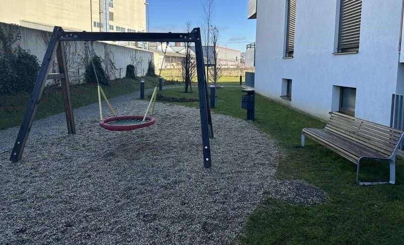 Kleiner Spielplatz mit Schaukel und Sitzbank in einem begrünten Außenbereich.