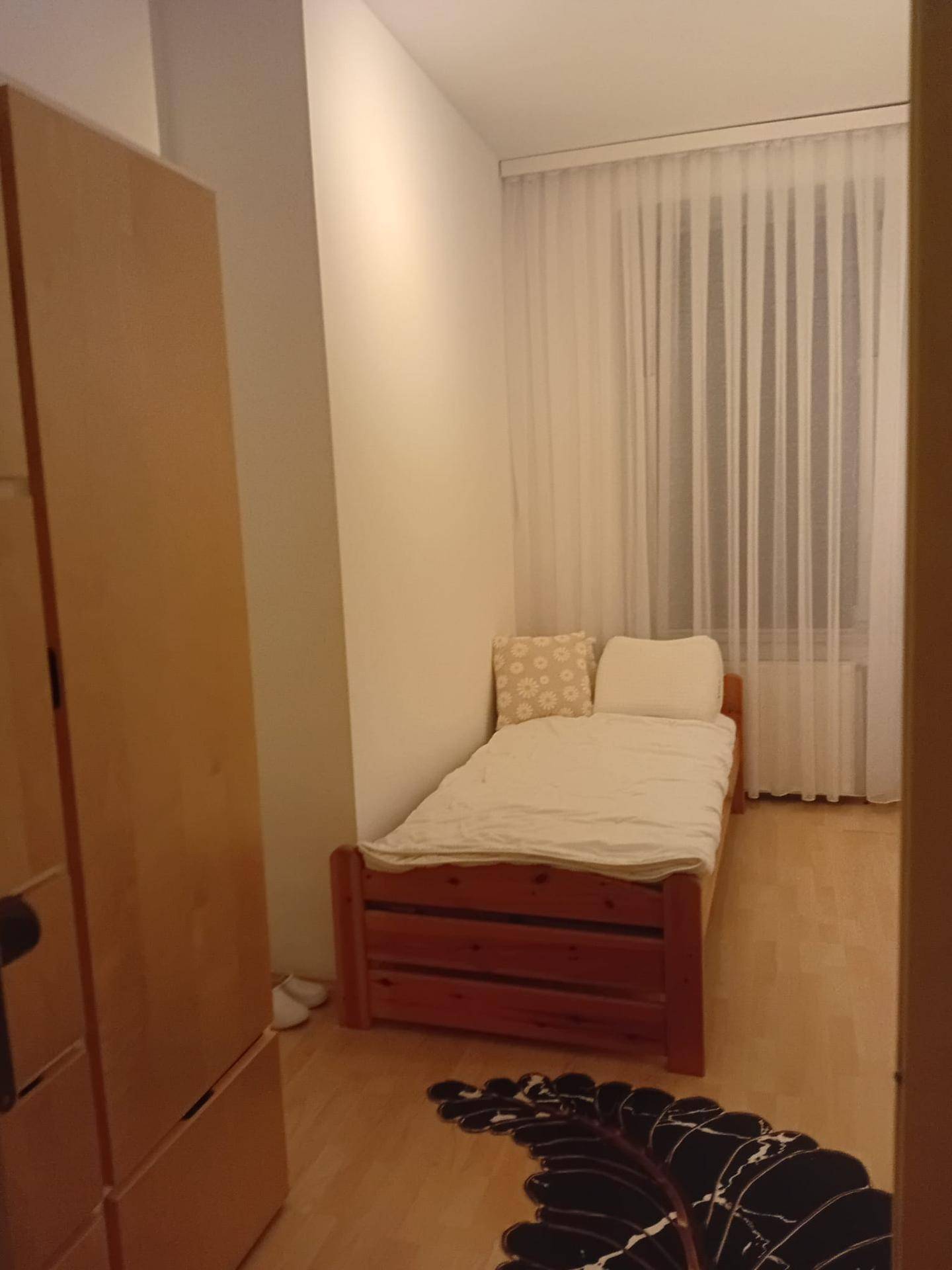 Schlafzimmer mit Einzelbett, hellem Holzschrank und einem Fenster mit transparenten Vorhängen.