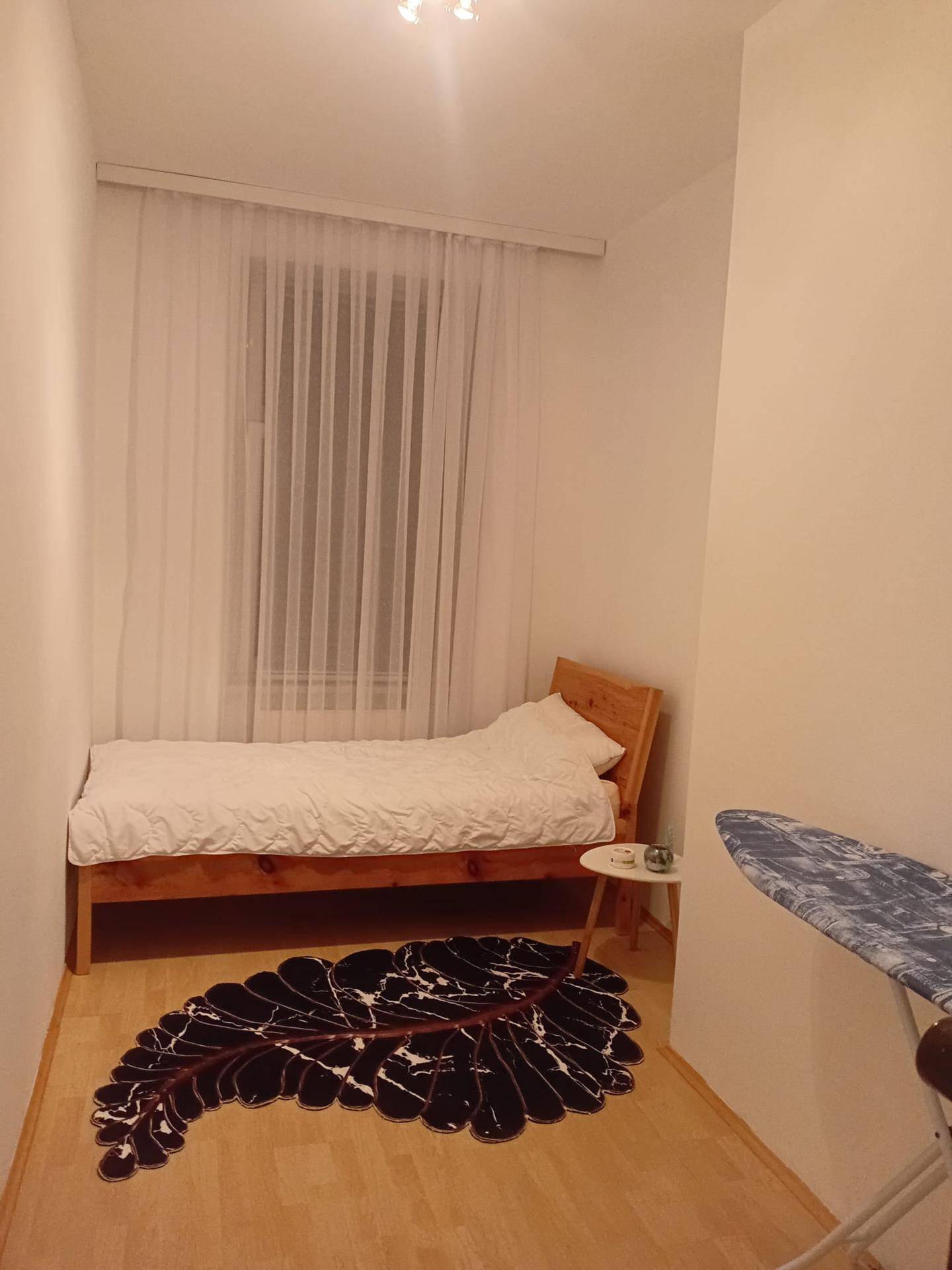 Kleines Schlafzimmer mit Einzelbett, hellem Holzrahmen und einem Fenster mit Vorhängen.