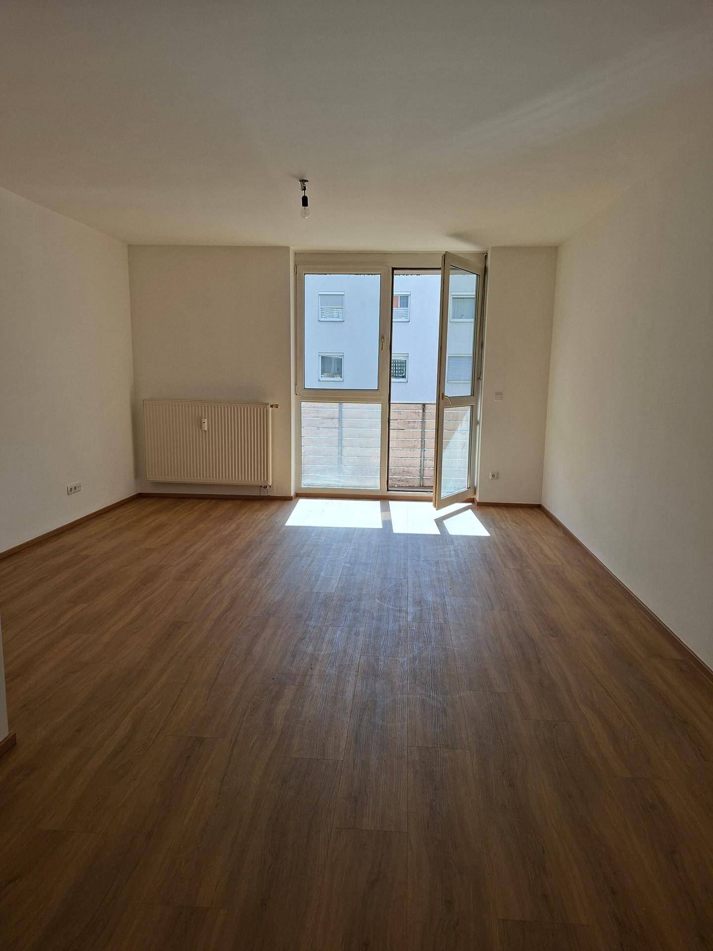 Lichtdurchfluteter Raum mit Laminatboden, Heizkörper und Balkonzugang.