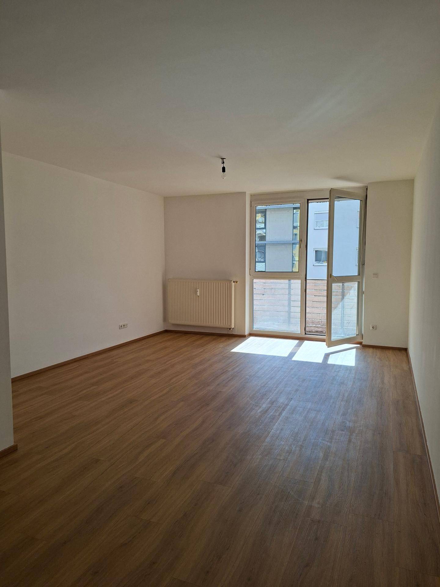 Heller Wohnraum mit Laminatboden, Heizkörper und großem Fenster zum Balkon.