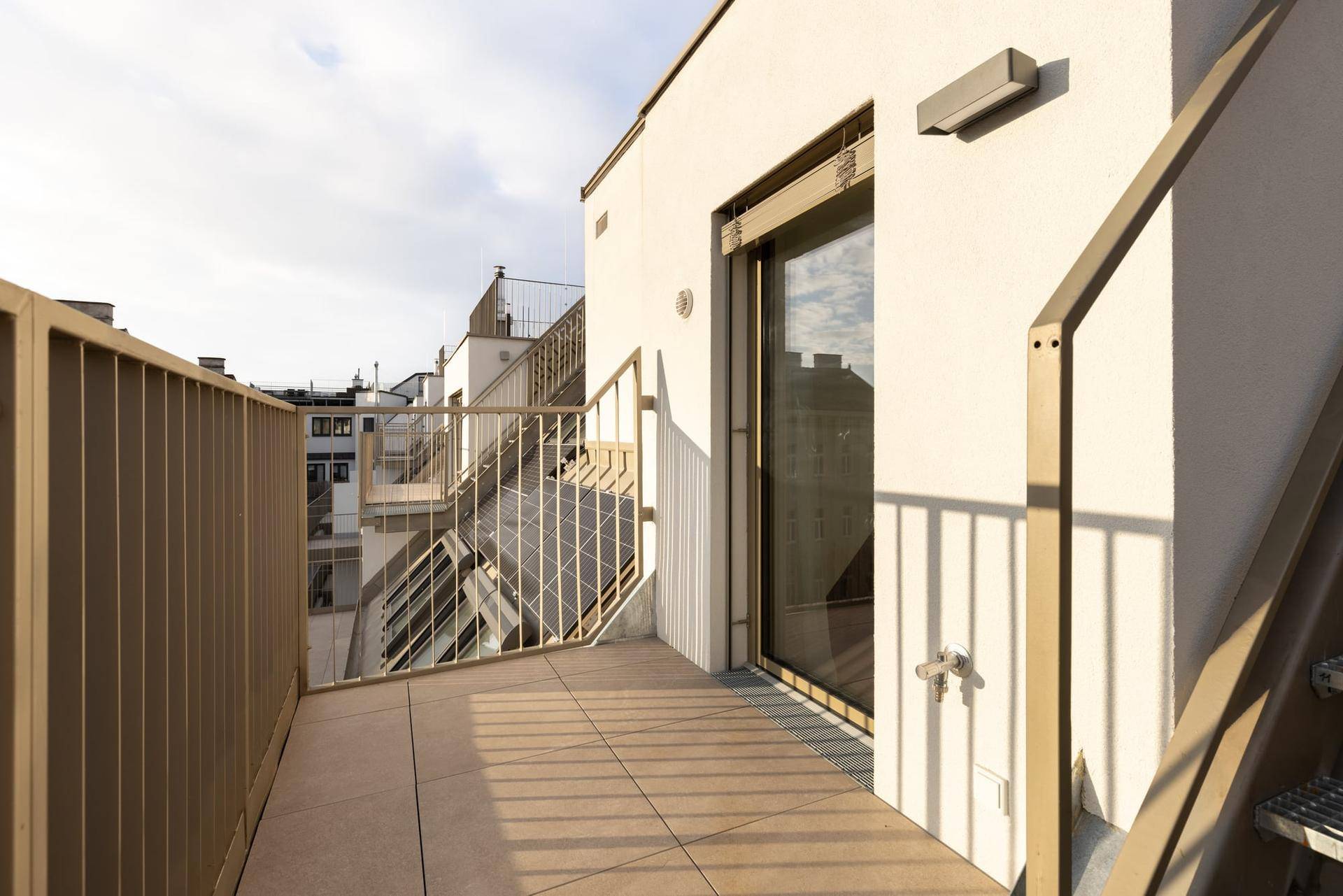 Sonniger Außenbereich mit Fliesenboden und moderner Metallbrüstung, zugänglich durch eine Glasschiebetür.