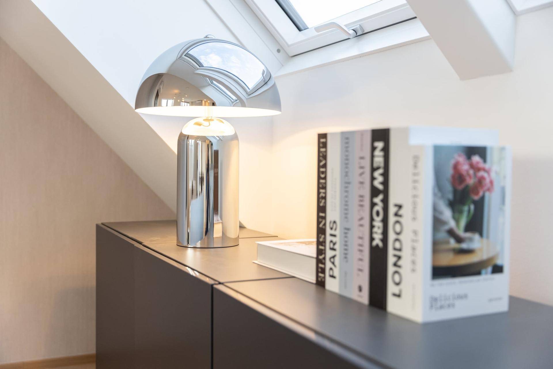 Detailaufnahme einer modernen Chromlampe und dekorativer Bücher auf einem dunklen Sideboard.