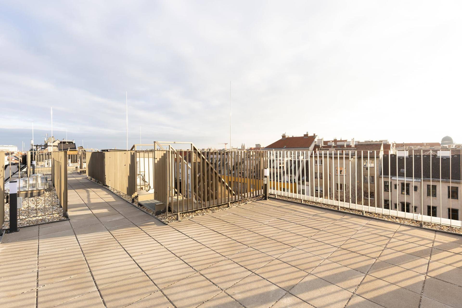 Geräumige Dachterrasse mit Fliesenboden und Metallgeländer, umgeben von städtischen Dächern.
